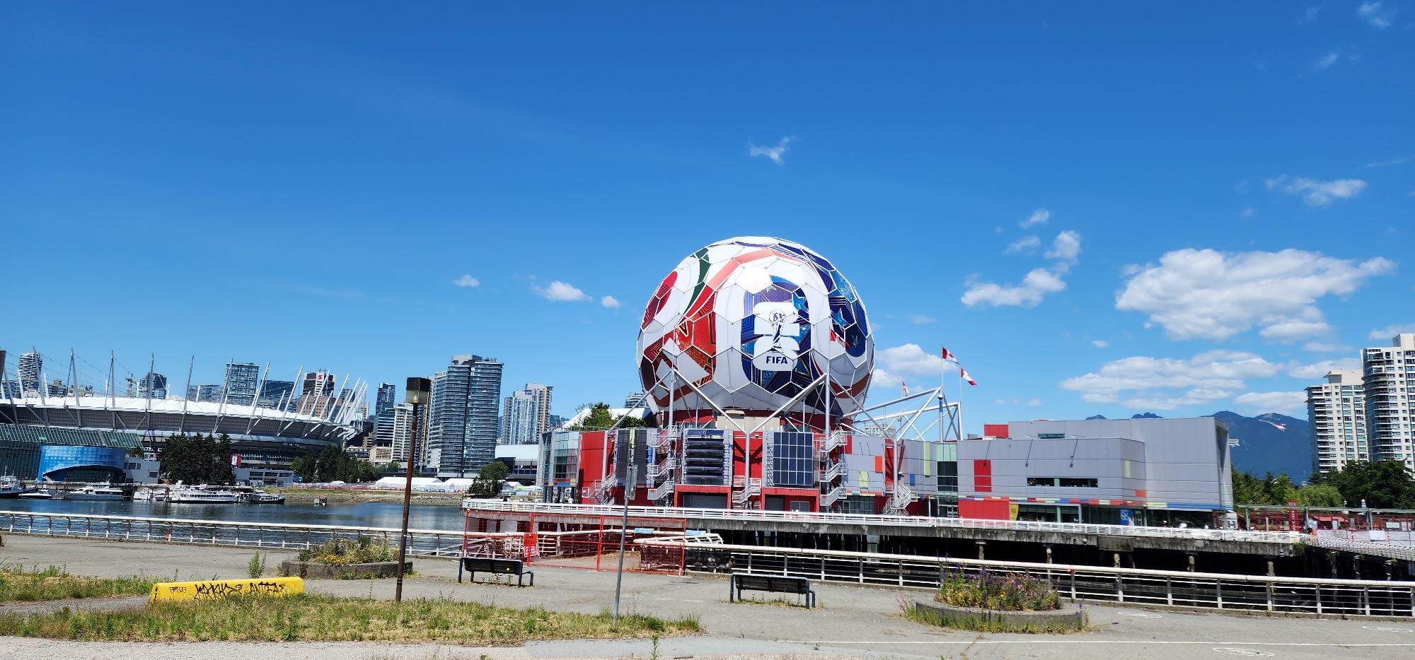 An outdoor, wide-angle shot captures a city waterfront scene on a clear, sunny day with a bright blue sky dotted with fluffy white clouds. In the foreground, a paved area with patchy grass and a yellow concrete barrier marked with graffiti is visible. Beyond this, a metal railing separates the foreground from a body of water, which reflects the sky. Several boats are docked on the water.
Dominating the mid-ground is a large, spherical structure resembling a soccer ball, emblazoned with the FIFA logo and colorful geometric patterns in red, white, and blue. This structure is mounted on a multi-level platform with red and grey exterior walls, visible stairs, and solar panels. To the left of the soccer ball structure, a large stadium with a distinctive white, cable-supported roof is partially visible, along with numerous tall city buildings in the background. Two Canadian flags fly from a pole near the soccer ball structure.
The background features a dense skyline of modern skyscrapers. In the far distance, mountains are visible, softened by a hazy blue atmosphere. The scene appears to be a public space with benches and a waterfront promenade, suggesting a location in a developed urban area with recreational amenities.