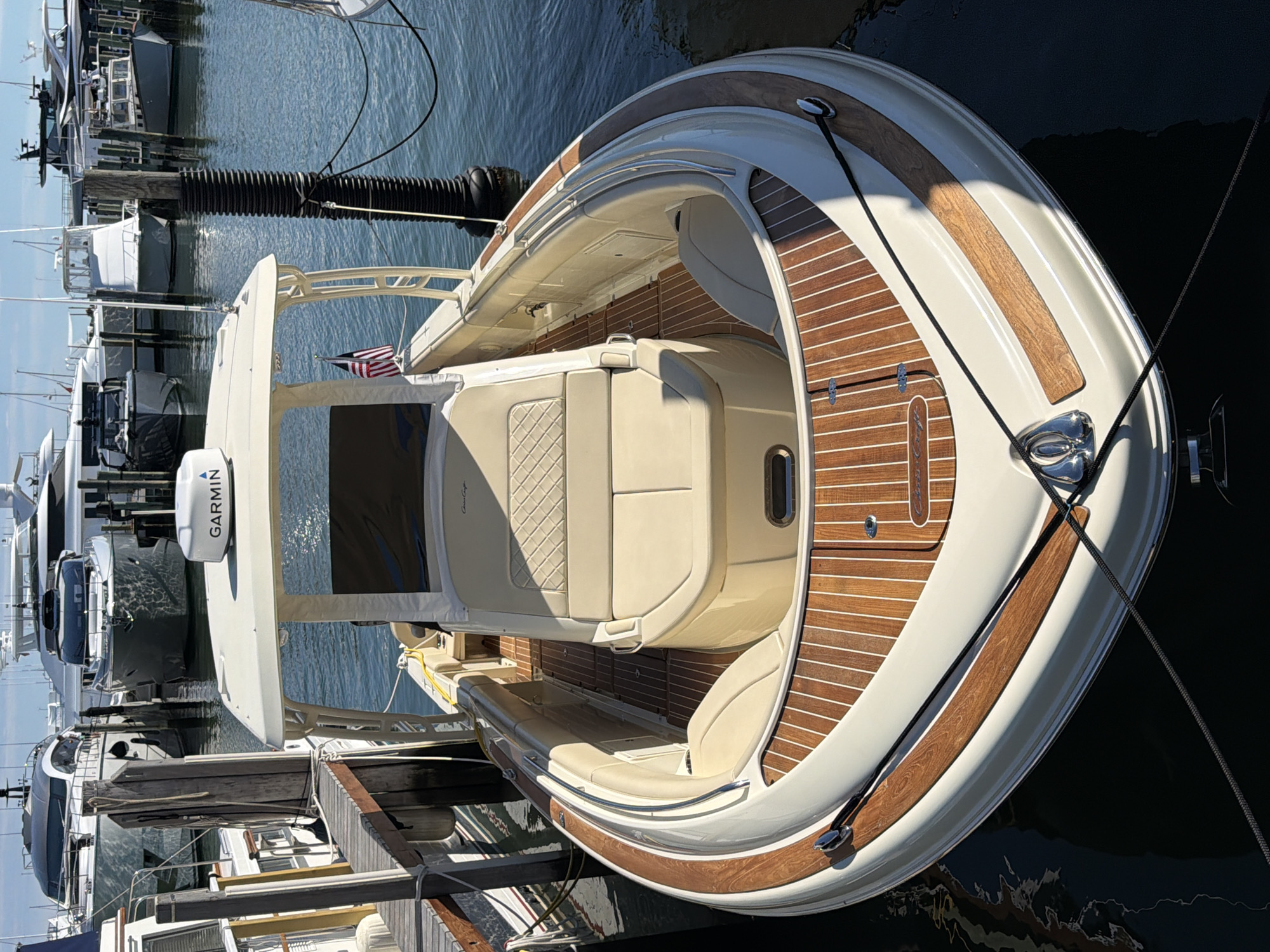 A white Chris-Craft boat is docked in the Village of Sag Harbor, USA. The boat is facing forward and is viewed from a slightly elevated angle. The interior of the boat features tan upholstered seating with diamond stitching on the backrests and a wood-planked floor. A Garmin GPS unit is visible on the white canopy above the helm. A small American flag is affixed to the starboard side of the boat. The boat is tied to a black mooring post with black ropes. In the background, other boats and yachts are docked in the harbor. The water is calm and reflects the sky and surrounding boats. The scene appears to be a sunny day.