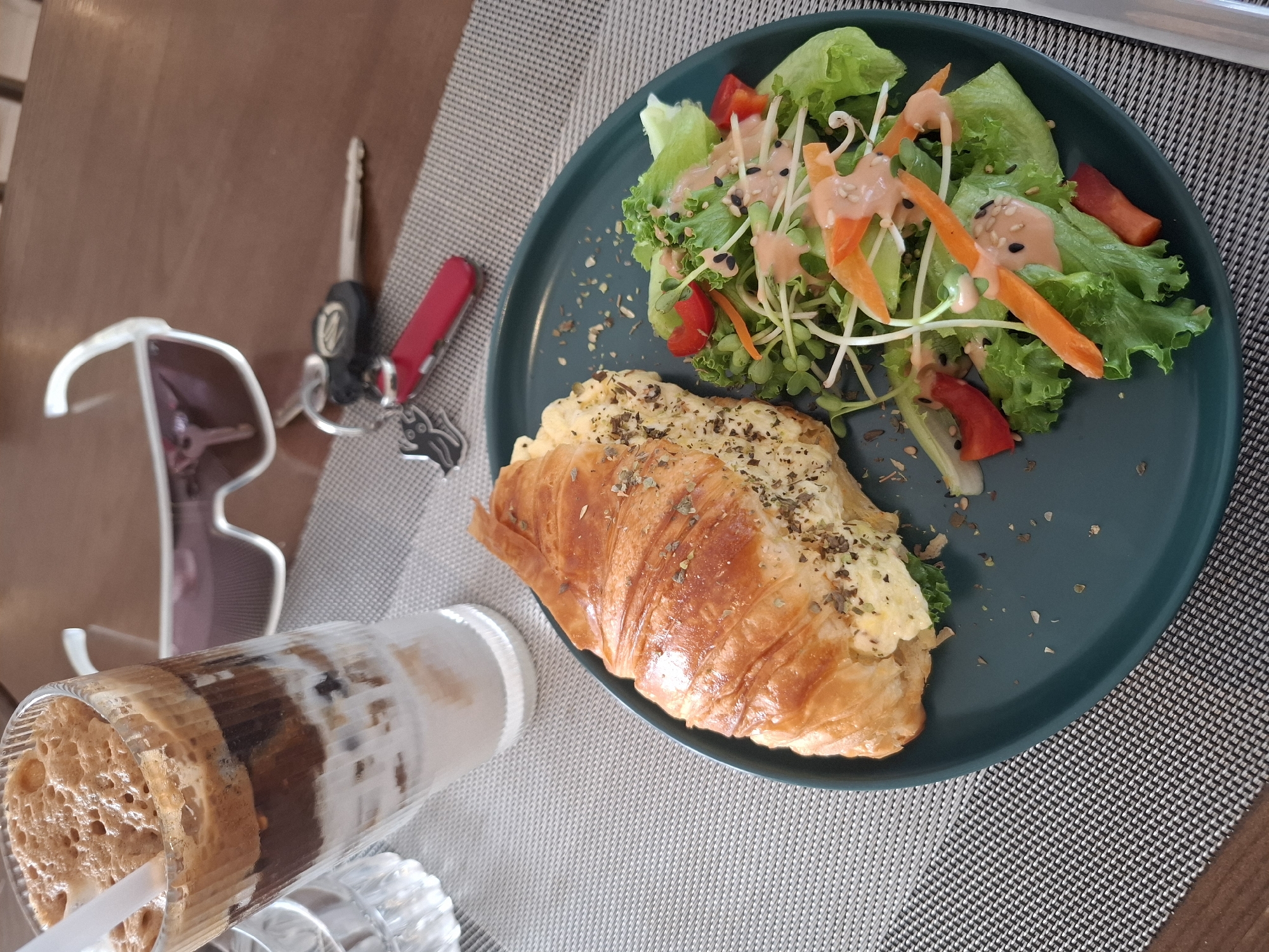 A high-angle, close-up shot shows a meal of a croissant and salad on a dark green plate. The croissant, halved lengthwise, is filled with scrambled eggs and herbs, and the salad consists of lettuce, shredded carrots, sprouts, and red bell pepper, all drizzled with a pinkish dressing and sprinkled with black sesame seeds. The plate sits on a grey placemat on a wooden table. To the left of the plate is a tall glass filled with a layered beverage, appearing to be iced coffee with a foamy top and a straw. Also on the table, near the top left, are a set of keys with a red Swiss Army knife attached. A pair of sunglasses with white frames rests on the table in front of the keys. The lighting suggests it is daytime, possibly morning or afternoon. The scene is set at a cafe or restaurant. The presence of sunglasses and keys could imply the diner is on the go or has just arrived. There is no visible text in the image.