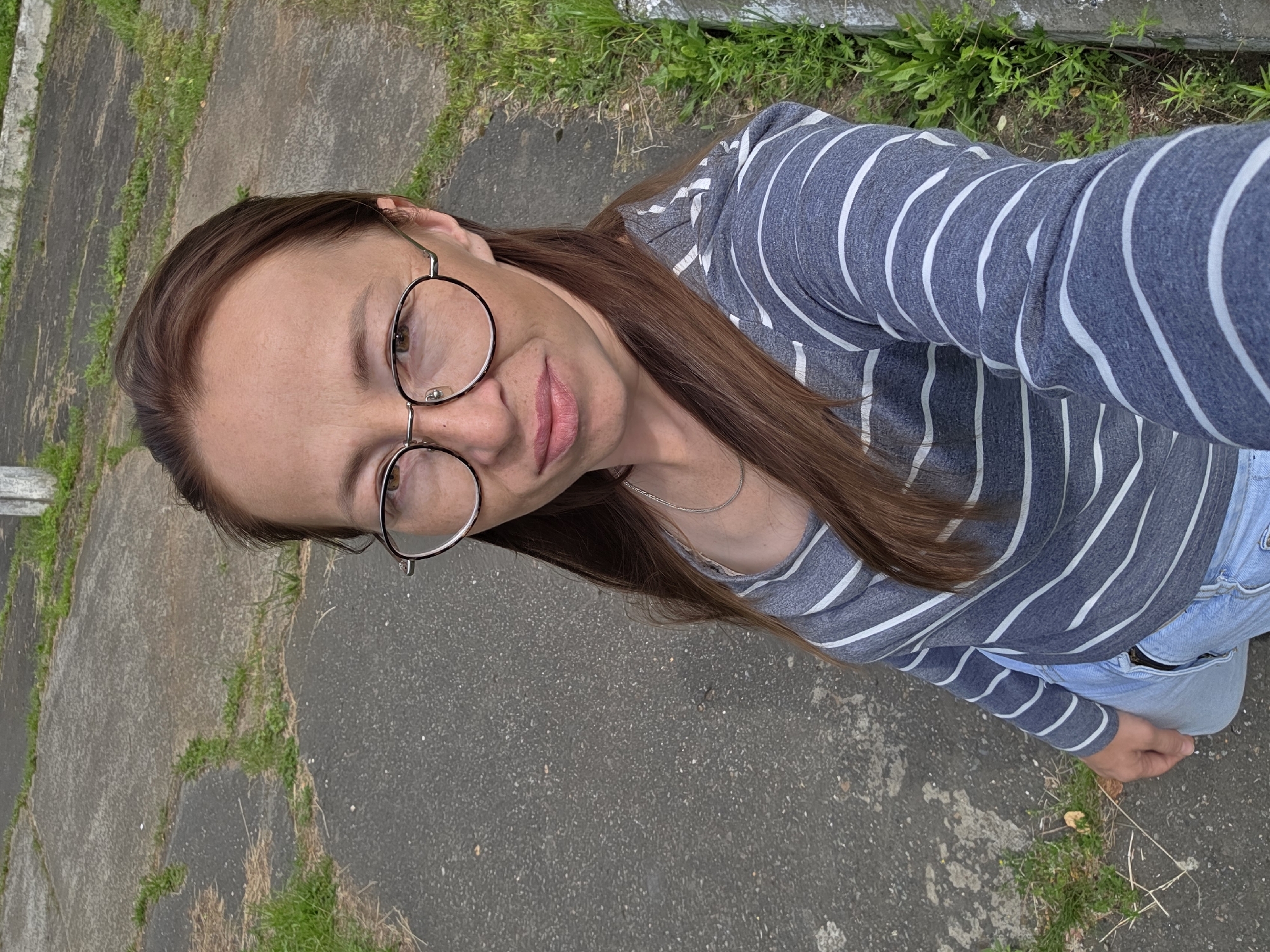 A woman with long brown hair and glasses is taking a selfie outdoors. She is wearing a grey and white striped long-sleeved shirt and light blue jeans. She is standing on a paved surface with patches of grass and some concrete structures visible. The location is the city of Chaikovsky, Russia.