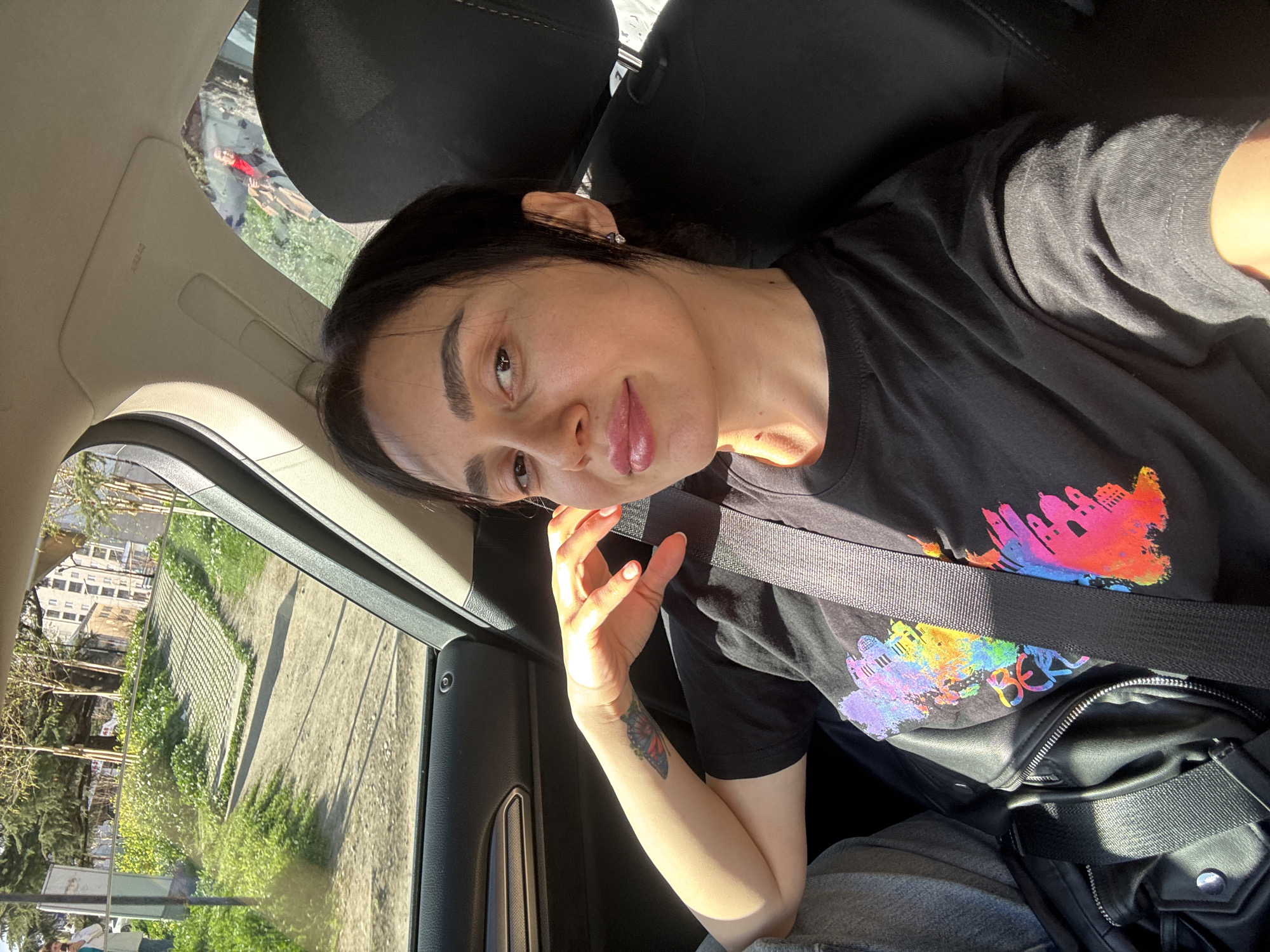 A woman is taking a selfie in the passenger seat of a car. She has dark hair pulled back, is wearing a black T-shirt with a colorful design, and has a seatbelt on. She's wearing a black crossbody bag. Outside the car window, there's a view of a sidewalk, grass, trees, and buildings. It appears to be Tbilisi, Georgia, due to the architectural style and general appearance of the buildings outside the window. The light suggests it's daytime. A person is visible in the car's sunroof.