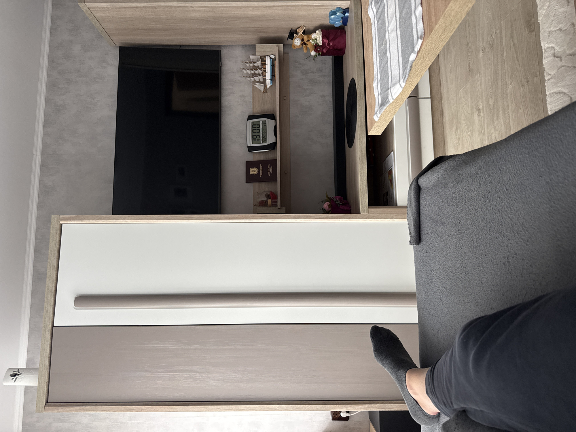 A sideways shot depicts a person seated, possibly on a sofa, in a room featuring a modern entertainment center. The person is wearing dark gray pants and one gray sock.
The entertainment center has a wooden top, a large black television, decorative items on shelves including a model ship, a digital clock displaying "19:00," and other small objects. The lower section of the unit has a white upper drawer and a beige lower drawer with a silver handle. An air freshener is attached to the side.
The sofa, covered in dark gray fabric, is partially visible.
The image was taken indoors. The location in the city of Lovech, Bulgaria is not discernible from the visual elements in the photograph.