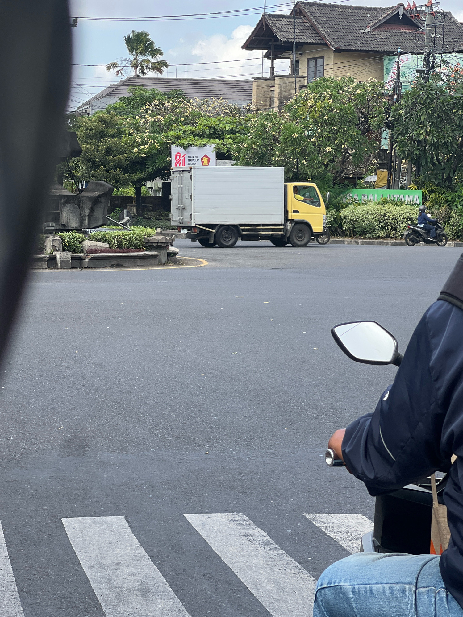 A yellow truck with a white cargo box is driving on a paved road in Denpasar, Indonesia. A person is riding a motorcycle in the background, and a crosswalk is visible in the foreground. Trees and buildings are also present in the scene.