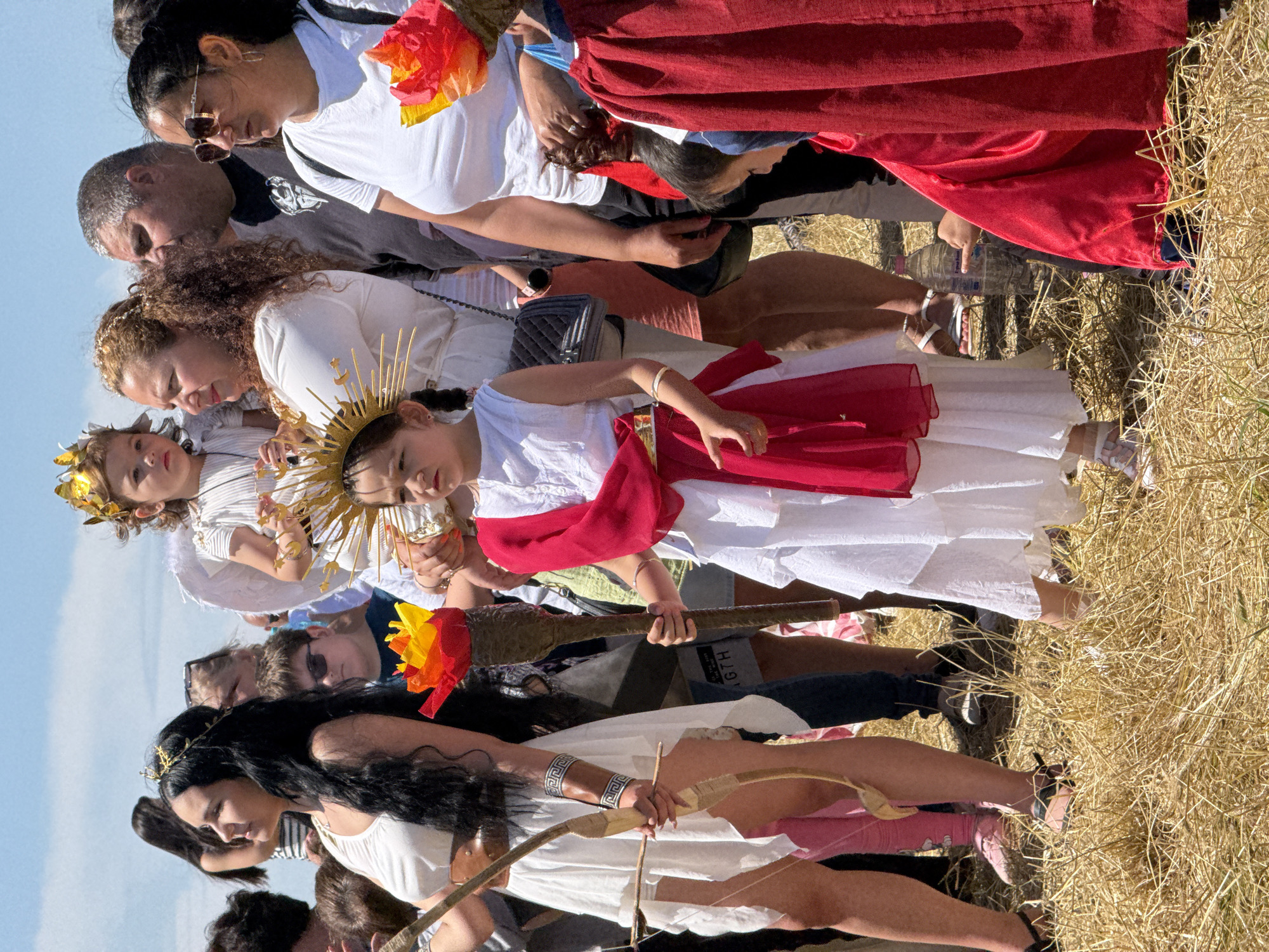 The scene depicts an outdoor gathering of individuals under a clear, light blue sky, taking place in the city of Gigen, Bulgaria. The ground is covered with dry, light brown, straw-like grass, illuminated by bright sunlight.
In the left foreground, a young woman with long, dark hair wears a white, sleeveless, draped dress with a brown belt and a golden, wreath-like headband. She holds a small, light-colored wooden bow with an arrow nocked, pointing slightly downwards. A patterned armlet adorns her right forearm. Her expression is neutral, and she gazes to the left.
Central to the image, a young child, possibly a girl, stands wearing a white layered dress with a prominent red sash or drapery over her left shoulder and waist. She wears a golden, spiky sun-ray crown on her head and holds a decorative torch with a red and yellow flame-like top in her right hand. Her gaze is directed straight ahead.
Immediately behind this child, an adult woman with curly brown hair, dressed in a white long-sleeved garment, holds a smaller child on her left hip. This younger child wears a white outfit with small white angel wings attached to its back and a golden crown on its head, looking towards the right.
To the right of the central group, an adult man with short, dark hair and a beard, wearing a dark t-shirt, stands looking forward. Adjacent to him, an adult woman with dark hair and sunglasses wears a white t-shirt and holds a large red and yellow flame-like prop similar to the torch. Her attention is directed towards the right.
Partial views of other individuals are visible throughout the background, including adults and children in various attire, some in casual wear. In the lower right foreground, a child's hand points towards a clear plastic water bottle with a pink cap lying on the dry grass.