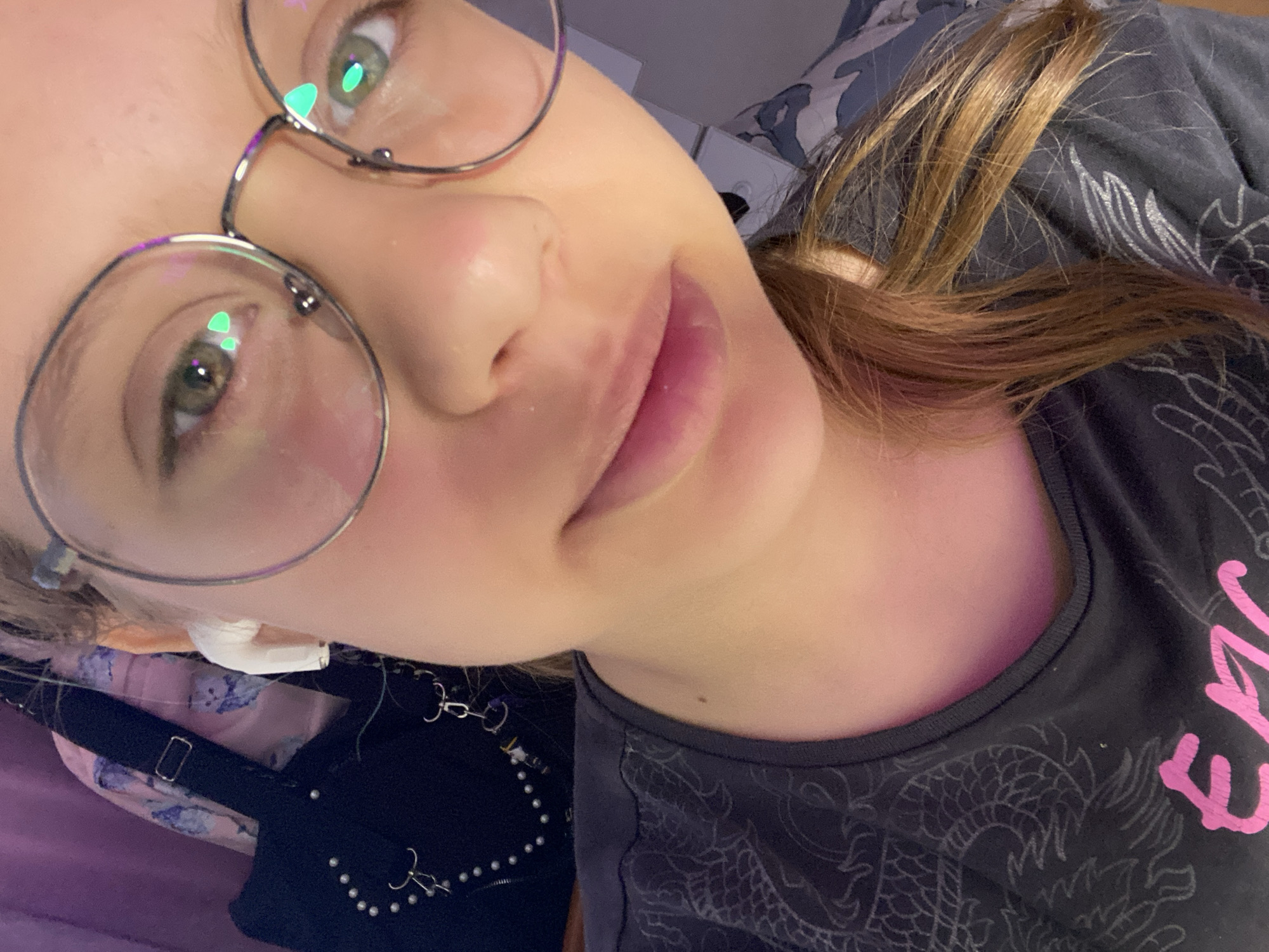 A close-up selfie taken indoors shows a young person with light brown hair, wearing large round glasses and white earbuds. They are wearing a dark gray t-shirt with a dragon design and pink lettering. The background includes a blurred image of a dark bag with beaded embellishments and a light pink garment with a floral pattern, suggesting a bedroom or personal space. The lighting is soft, with reflections visible on the glasses. The image does not contain any information about the city of Zassovskaya, Russia, or any specific location.