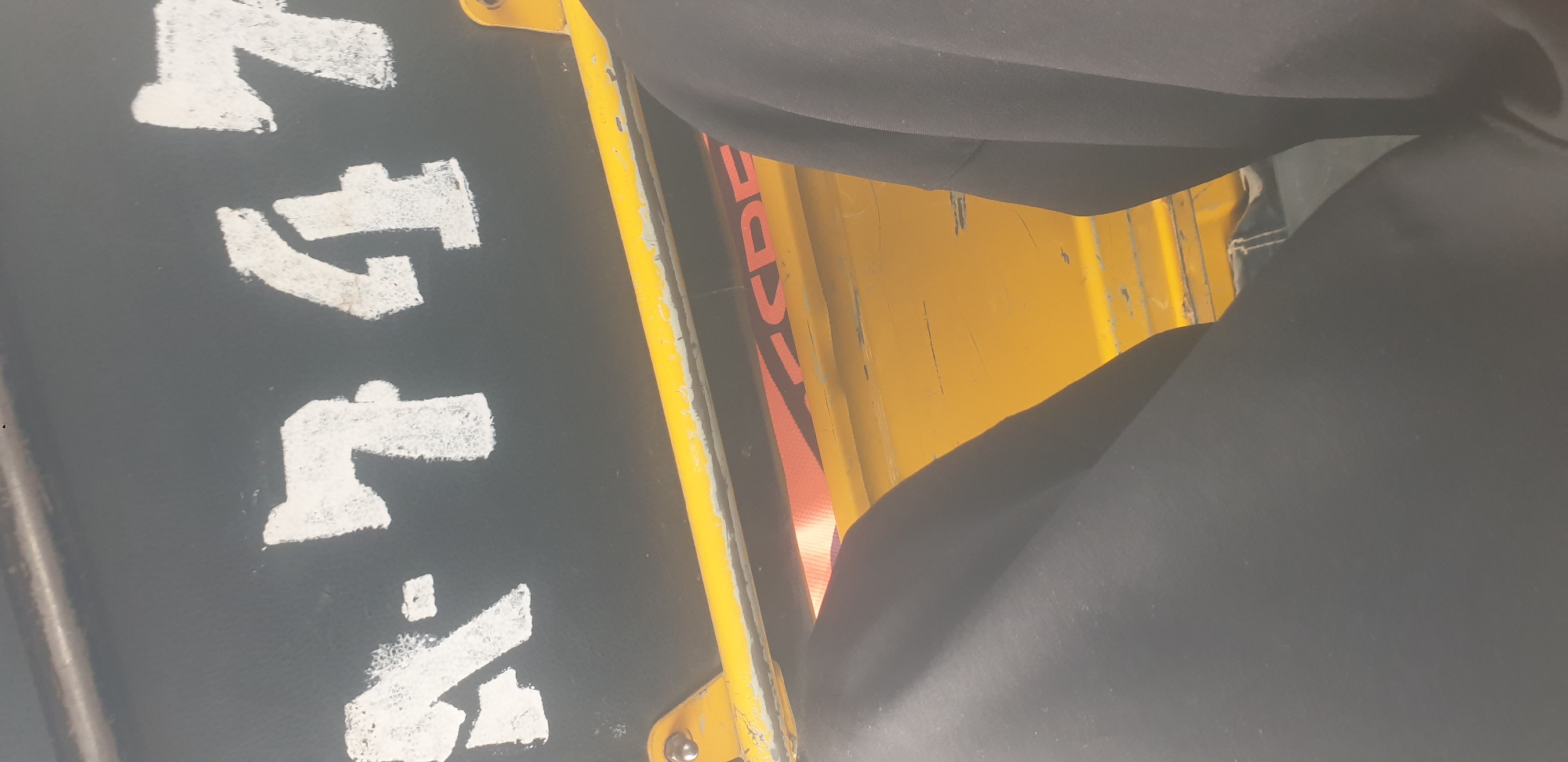 The image shows a close-up of what appears to be the side of a vehicle, possibly a truck or bus, in Barakin Lerawi, Nigeria. White lettering on a black background reads "N-747". Below this, a yellow metal bar runs horizontally across the image, showing signs of wear and tear. Part of a person's dark clothing is visible in the foreground on the left and right, obscuring a portion of the yellow bar and the vehicle's interior. The lighting suggests it is daytime.