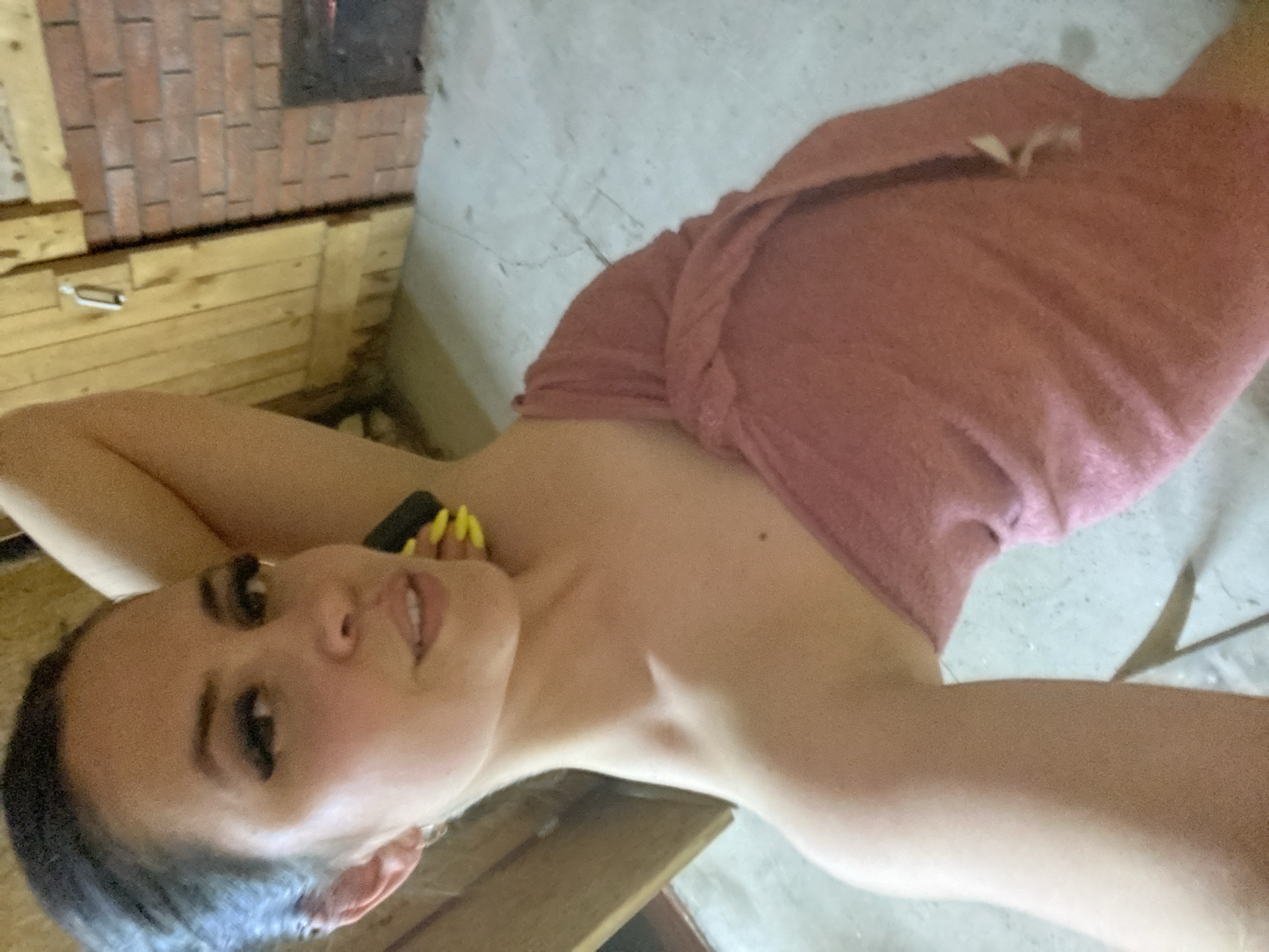 A fair-skinned woman is lying down, partially wrapped in a pink towel. She has dark hair and is wearing noticeable false eyelashes. Her left arm is raised above her head, revealing painted yellow fingernails. The background includes a brick and wood structure, possibly a sauna or rustic building. A concrete or stone surface is also visible in the background. The exact location in Zhigalki, Russia cannot be definitively determined from the image.