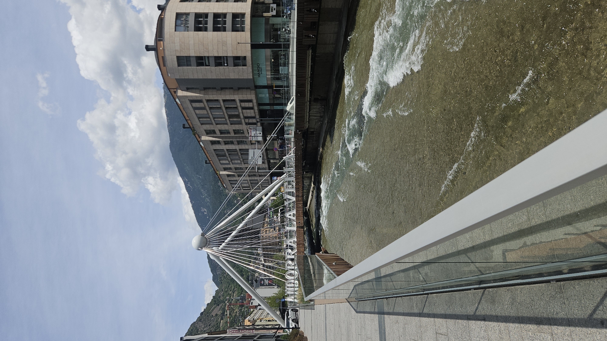 This is a daylight, eye-level shot of the Pont de Paris bridge spanning the Valira River in Andorra la Vella, Andorra. The image is taken from a paved pedestrian walkway on the near bank, looking across the river towards the city center and surrounding mountains.
In the immediate foreground, a modern railing with a light gray top rail and clear glass panels runs diagonally from the lower left to the mid-right. The walkway is paved with light-colored rectangular tiles, and shadows are cast from the railing onto the pavement on the left.
The Pont de Paris, a contemporary cable-stayed bridge, is the central feature, characterized by a prominent white spherical mast from which numerous white cables fan out to support the bridge deck. Large, white, capital letters spelling "ANDORRA LA VELLA" are mounted horizontally on the bridge structure, facing the viewer.
Beneath the bridge, the Valira River flows from left to right, exhibiting visible rapids and white foamy crests on its brownish-green surface, suggesting a shallow, rocky bed. The riverbanks are reinforced with concrete and wooden structures.
On the far side of the river, multi-story modern buildings with light-colored stone or concrete facades line the street. One building on the right features a distinctive curved corner, with large ground-floor windows displaying a "MANGO OUTLET" sign. Traffic signs are visible on poles near the buildings on the far side of the bridge.
In the mid-ground, a white SUV is partially visible on the left side of the bridge's road, and other vehicles can be seen further down the street on the right.
The background is dominated by steep, green, tree-covered mountains, rising under a bright blue sky dotted with scattered white cumulus clouds. No identifiable people are clearly visible in the scene.