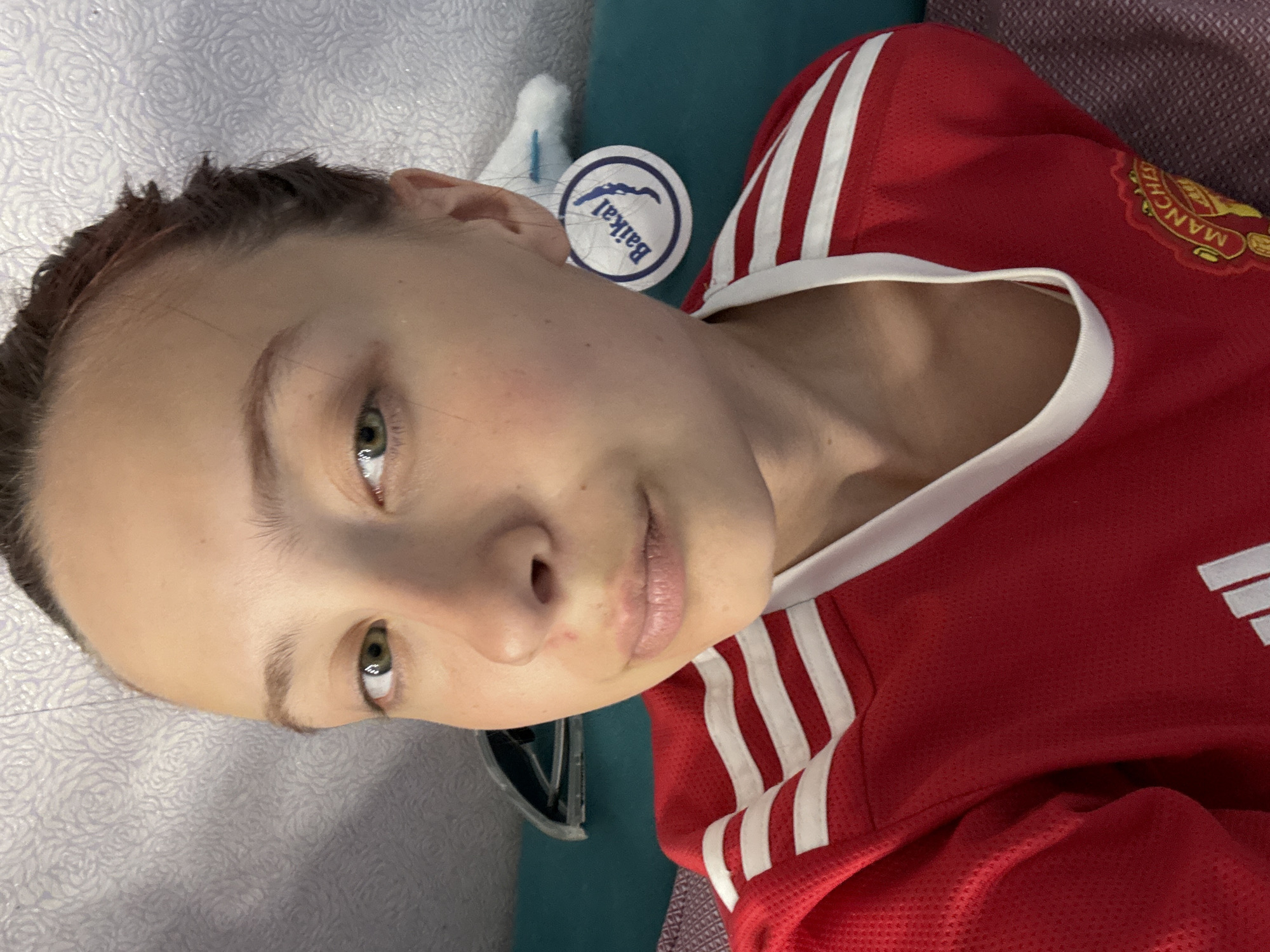 A young woman with fair skin and dark hair, appears to be lying down. She is wearing a red athletic jersey with white stripes, featuring a sports team logo on the right side of the chest. There's a round sticker with the word "Baikal" on it, positioned near her ear. The background includes elements of interior decor, with a patterned, likely textured, surface visible. The location is Прикамский, Russia.