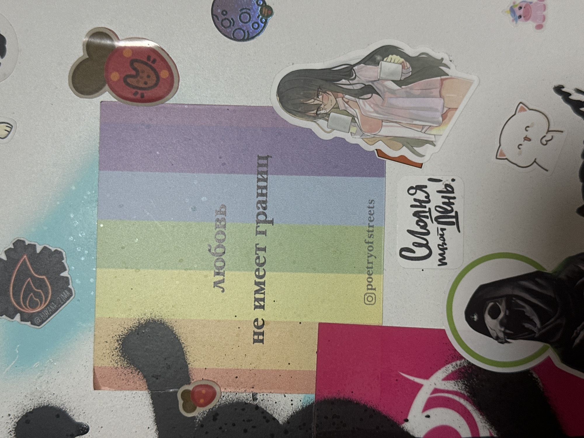 The image displays a collage of stickers on a white, textured surface, possibly a wall or a notebook. The central element is a square sticker with a rainbow gradient, featuring Russian text that translates to "love has no borders." Below this text, there is a small Instagram handle "@poetryofstreets." To the right of the rainbow sticker is a sticker of an anime-style girl with long dark hair, wearing glasses and a pink outfit, holding two white cups. Above her is a round, holographic sticker with concentric circles. To the left of the rainbow sticker, there is a large black spray-painted graphic with a pink spray-painted section at the bottom left. Several smaller stickers are also present, including a strawberry-like sticker with a face, a sticker with a cat-like silhouette and glowing red eyes, a sticker of a stylized skull wearing a hood, a small sticker with a white cat, and a pink unicorn sticker. The overall scene appears to be a personal decorative arrangement, possibly in an urban environment, but no specific location in Moscow, Russia, can be determined from the visual information provided. The lighting suggests a well-lit indoor or overcast outdoor setting.