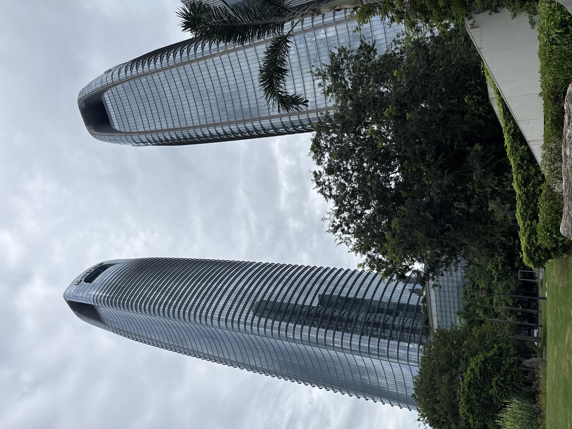 This image shows two modern skyscrapers in Tianhe District, China, under a cloudy sky. The buildings are tall and sleek, with curved, glass facades that reflect the overcast sky. The skyscraper on the left has a distinct, almost oval-shaped opening at its peak. The skyscraper on the right is slightly shorter and also has a curved profile. In the foreground, there are lush green trees and bushes, with a patch of grass to the left. Part of a concrete structure with layered greenery is visible in the lower right quadrant. The scene suggests an urban park or plaza adjacent to these office buildings.