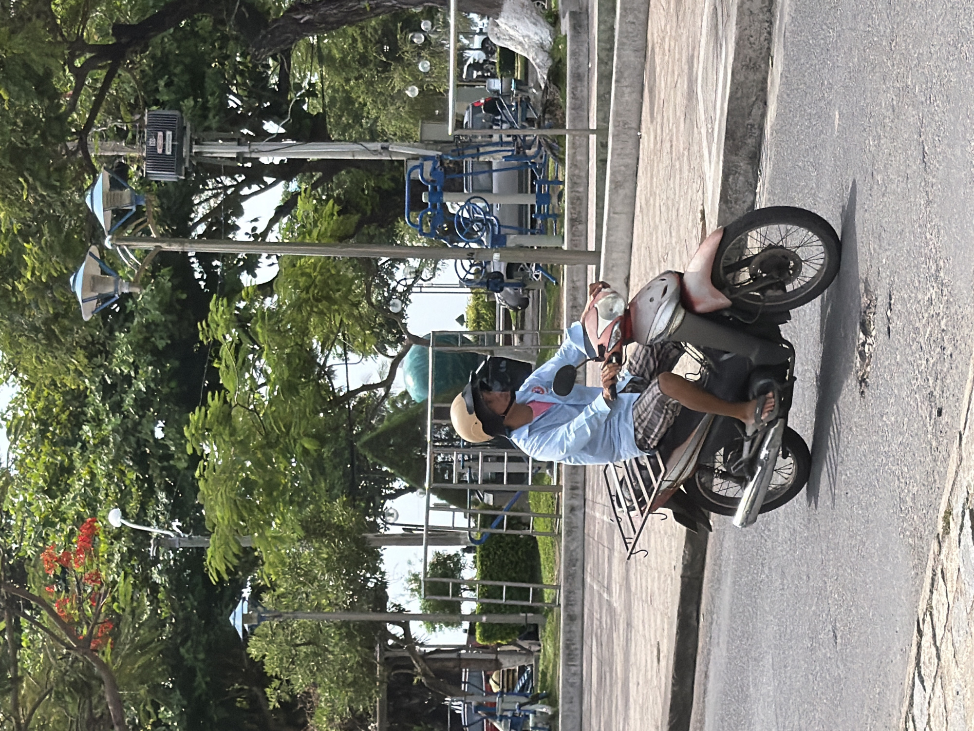 A person is riding a scooter down a paved street in Nha Trang, Vietnam. The person is wearing a light blue long-sleeved shirt, plaid shorts, flip-flops, and a light brown helmet with a dark visor. The scooter is red and silver, with a metal rack on the back. The street is lined with trees and a sidewalk. In the background, there are some outdoor exercise machines and streetlights. The sky is cloudy.