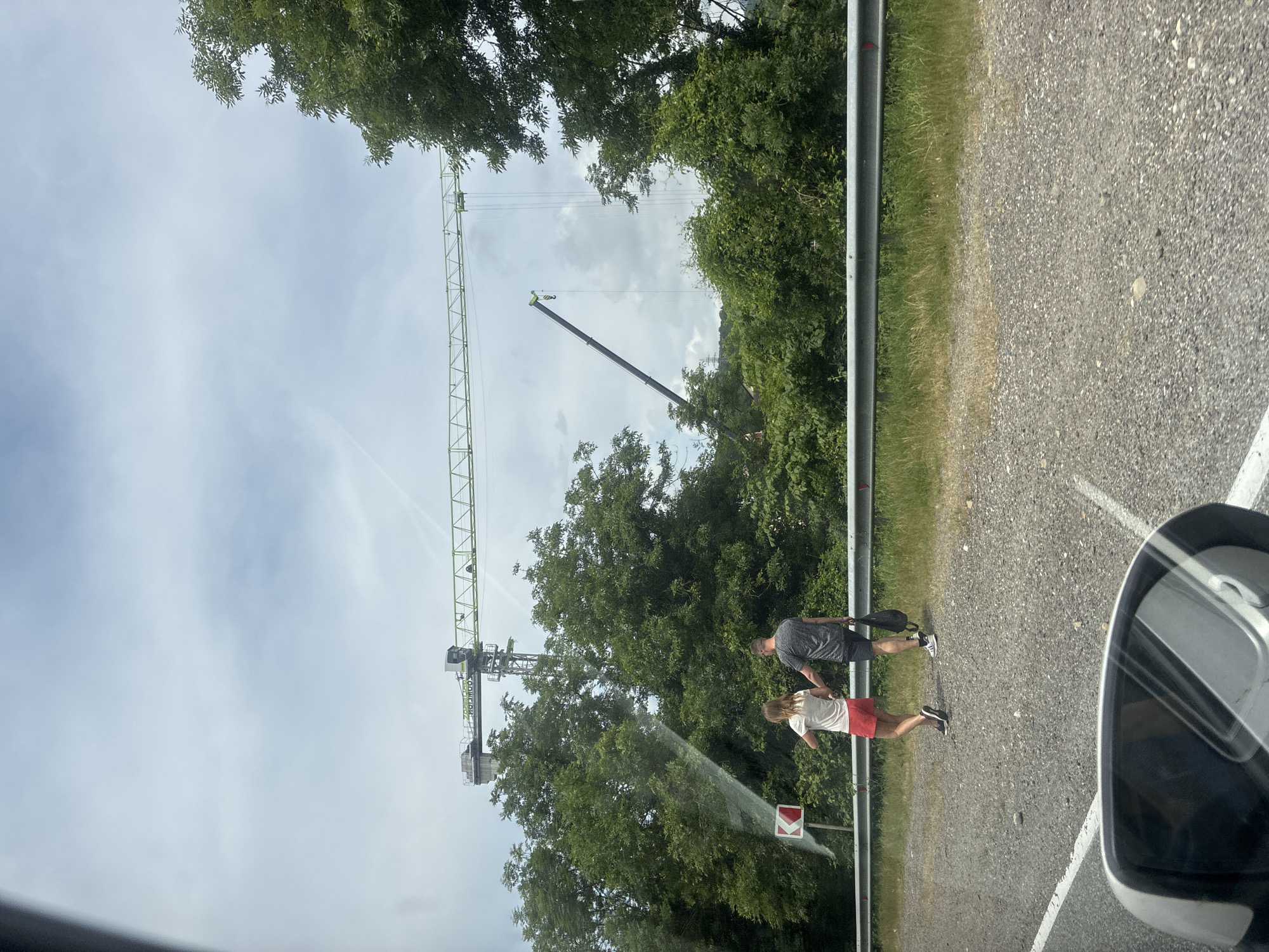 A couple walks hand-in-hand along a gravel shoulder next to a road, with a metal guardrail separating them from a grassy area and dense green trees. In the background, a large construction crane stands tall against a cloudy sky, with another, smaller crane arm extending from its side. The couple is walking away from the camera, the female wearing a white t-shirt and red shorts, and the male in a grey t-shirt and black shorts, carrying a black backpack. A directional sign indicating a curve to the left is visible near the couple. The scene appears to be on the outskirts of Novomikhailoskiy, Russia, as indicated by the context provided, amidst a predominantly natural environment with signs of ongoing construction.