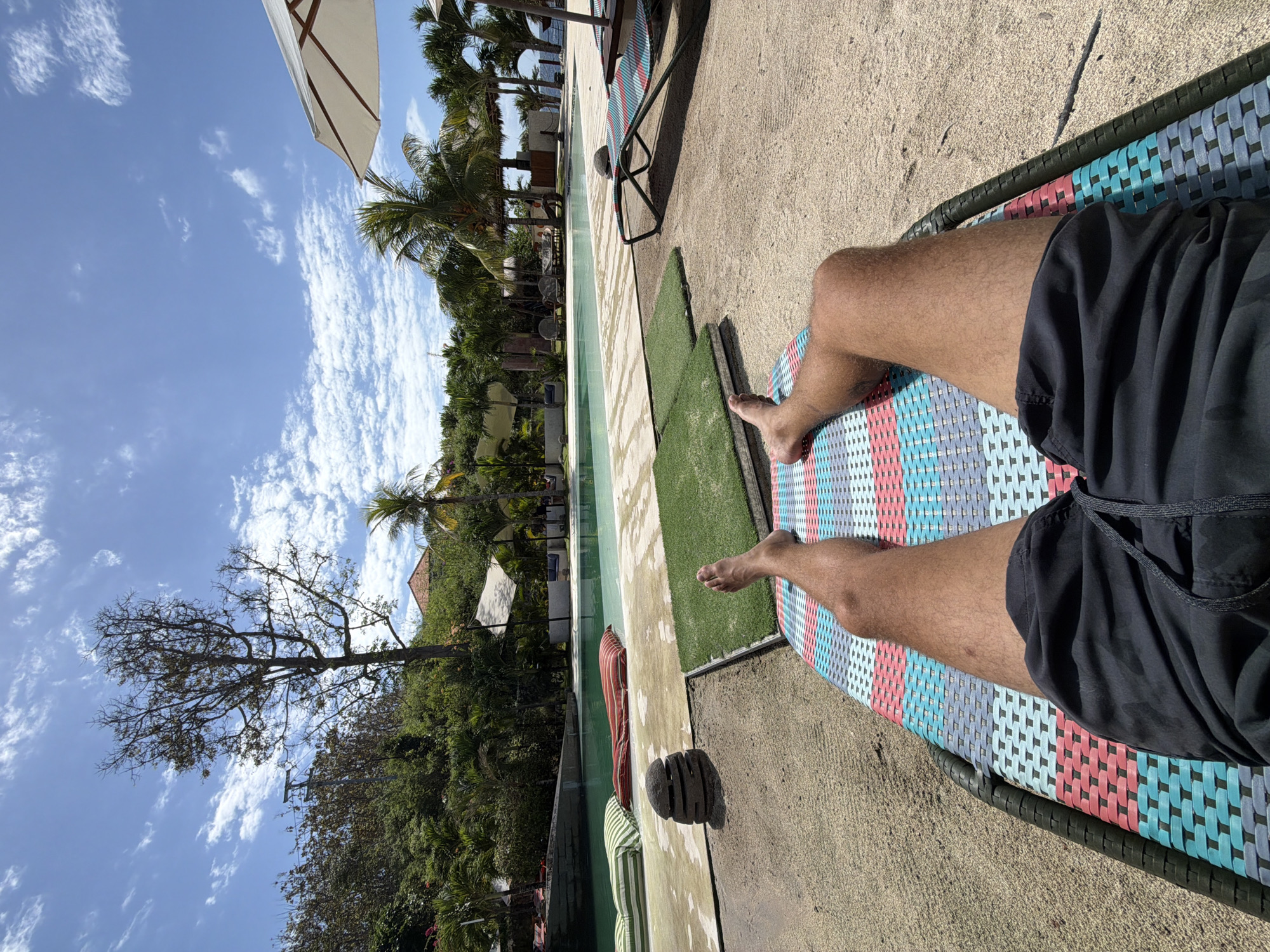 A first-person perspective shot captures a person relaxing on a multi-colored lounge chair. The person's legs, clad in dark swim trunks, are extended forward, their feet bare. The lounge chair rests on a sandy surface adjacent to a swimming pool. The pool, filled with clear water, is surrounded by a paved area with some decorative elements. Beyond the pool, lush green vegetation, including palm trees, forms a dense backdrop. The sky above is a bright blue, dotted with scattered white clouds, suggesting a sunny day. The scene is set outdoors, likely at a resort or a private villa in Nusa Penida, Indonesia, given the tropical foliage and swimming pool.