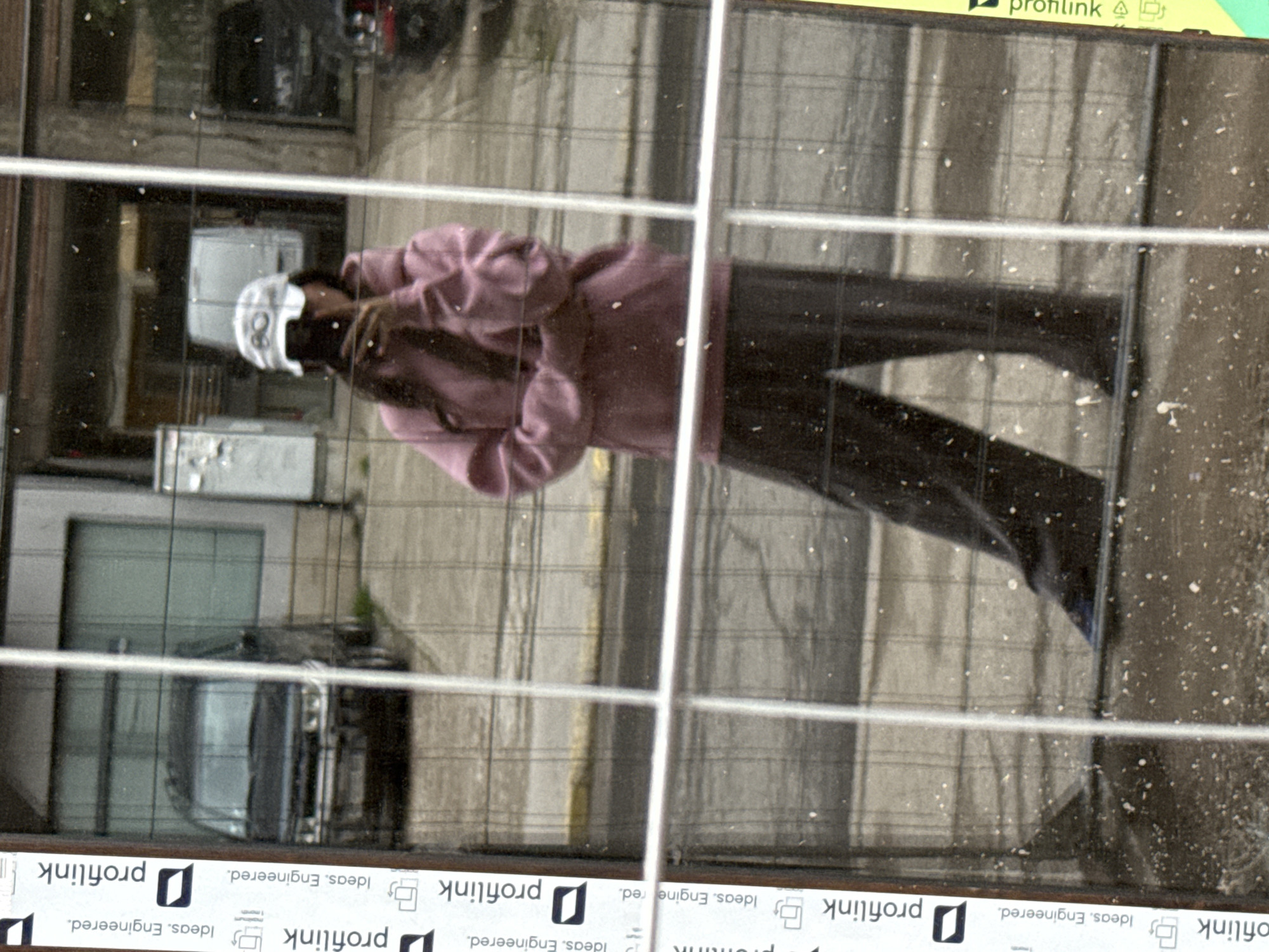 This media file presents an image rotated 90 degrees clockwise, primarily showing a reflection on a grimy glass surface. The central figure is a person reflected in the glass, appearing to wear a light purple or mauve long-sleeved top or jacket and dark trousers. Their head is covered by a white cap or bandana, partially obscuring their face.
The glass surface is marked by prominent white horizontal and vertical grid lines, dividing the view. It is also visibly dirty, covered with numerous small white specks. Through the reflection and possibly beyond the glass, a street scene is discernible, including parked vehicles such as a dark-colored car, and reflections of urban structures or paved ground.
Along the bottom and top edges of the rotated image, a yellow strip is visible containing text. The recurring text elements include "profilink" and "Ideas. Engineered." This scene is situated in Plovdiv, Bulgaria, and appears to be illuminated by natural daylight.