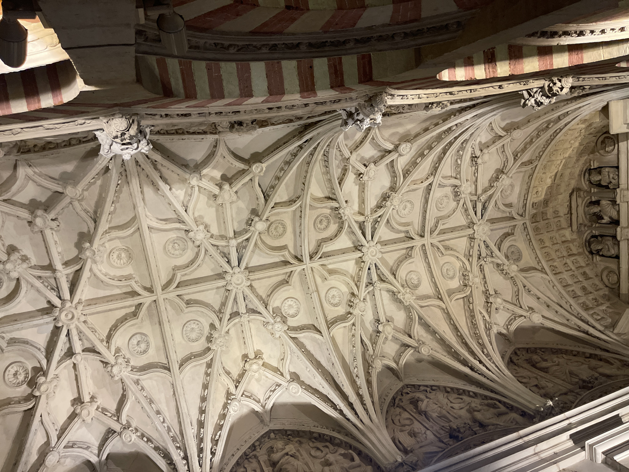 The image captures a low-angle view of the intricately decorated ceiling of the Mezquita-Cathedral of Córdoba, Spain. The ceiling is a stunning example of Gothic ribbed vaulting, characterized by its complex network of stone ribs that divide the ceiling into a series of panels. These panels are further adorned with detailed plasterwork, featuring rosettes and circular motifs. The ribbed vaulting extends across the vast space, creating a sense of height and grandeur.
To the right of the vaulted ceiling, a portion of a pillar is visible, distinguished by its alternating bands of red and white stripes, a characteristic architectural element of the Mezquita. Small, ornate corbels, sculpted with grotesque or animalistic figures, protrude from the junctions of the ribs and pillars, serving as decorative supports.
On the left side of the image, a section of a richly carved stone relief is partially visible. This relief appears to depict figures, suggesting historical or religious narratives embedded within the architecture.
The lighting in the image appears to be artificial, casting shadows that accentuate the three-dimensional qualities of the ceiling's ornamentation. The overall impression is one of historical depth and architectural artistry.