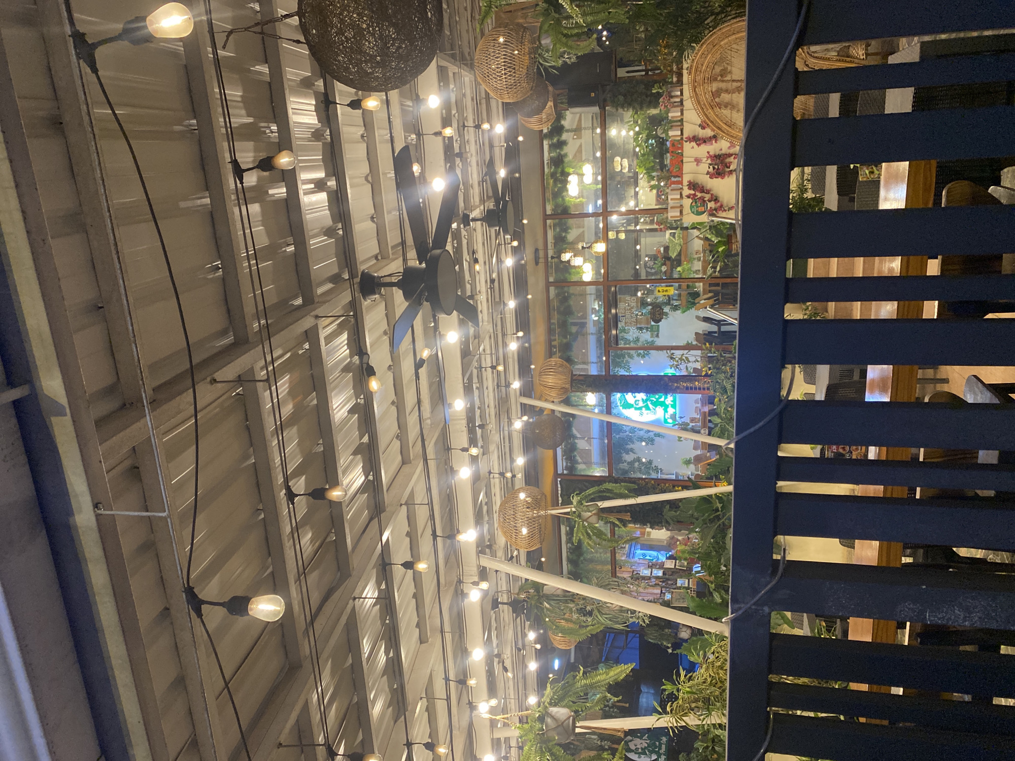 The image depicts the outdoor seating area of a cafe in Davao City, Philippines, illuminated by string lights and pendant lamps. Two large black ceiling fans are visible, along with several hanging baskets of greenery. Woven pendant lamps and decorative woven circular elements add to the ambiance. The cafe appears to have a rustic or bohemian aesthetic. Through the windows, various plants, tables, and chairs are visible, along with some signage, including one that appears to say "OPEN". The foreground features a dark-colored fence or railing, partially obscuring the view of the seating area. The overall scene suggests a relaxed, evening dining or gathering environment.