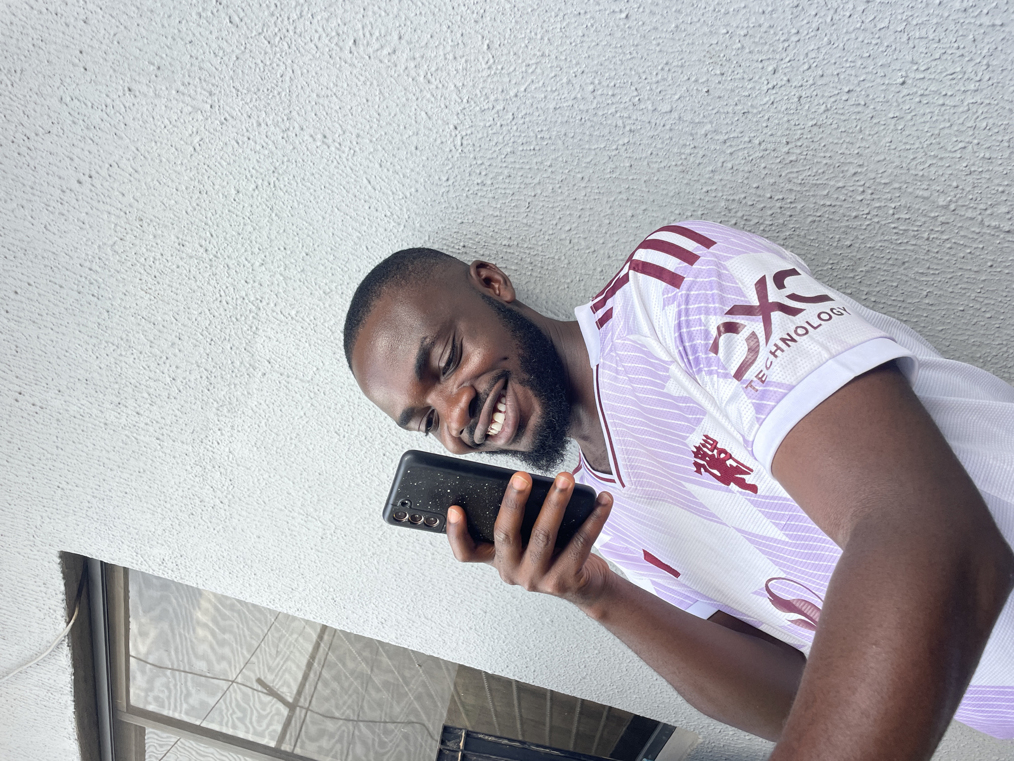 A Black man is taking a selfie with his phone, leaning against a textured white wall. He is wearing a white jersey with light purple stripes and maroon accents. His phone is black with a distinctive camera array. To the left of the man, a window with screens is visible. The scene appears to be outdoors, possibly in a residential or commercial area of Jos, Nigeria, given the architectural style and natural lighting. The man is smiling broadly, indicating a positive mood. The overall impression is casual and candid.
