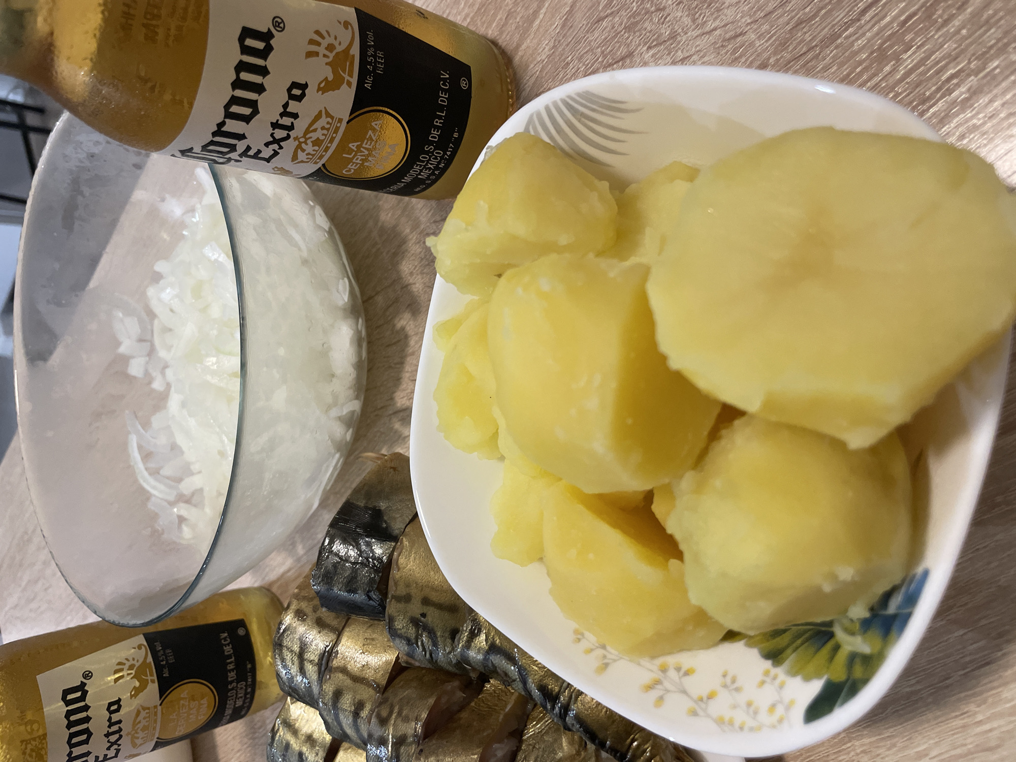 The image shows a table with food and drinks. There are two bottles of Corona Extra beer, a bowl of chopped onions, a dish of sliced smoked mackerel, and a bowl of boiled potatoes. The overall impression is of ingredients being prepared for a meal, possibly a picnic or outdoor gathering. The lighting is natural, suggesting it was taken during the day. There are no people visible in the image. The scene is set on a wooden table.
