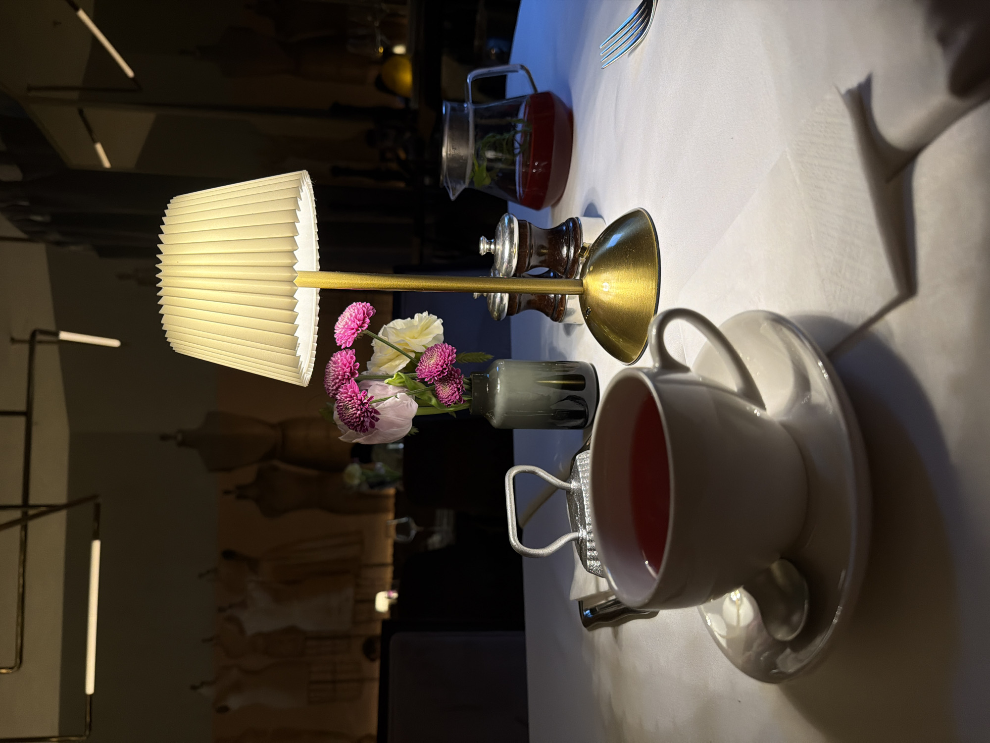 The image captures a table setting indoors. A white tablecloth is spread across the table on which a cup of tea or coffee sits on a saucer with a spoon. Next to the cup, there is a small vase of pink and white flowers. Adjacent to the flowers is a brass-colored lamp with a pleated lampshade, along with a glass pitcher filled with a red liquid and what appears to be mint leaves. A fork is present in the upper right corner of the frame. The background reveals a restaurant setting with decorative mannequins and linear lighting fixtures. Based on the scene’s style, it might suggest a location in a sophisticated urban area, potentially Moscow, Russia.