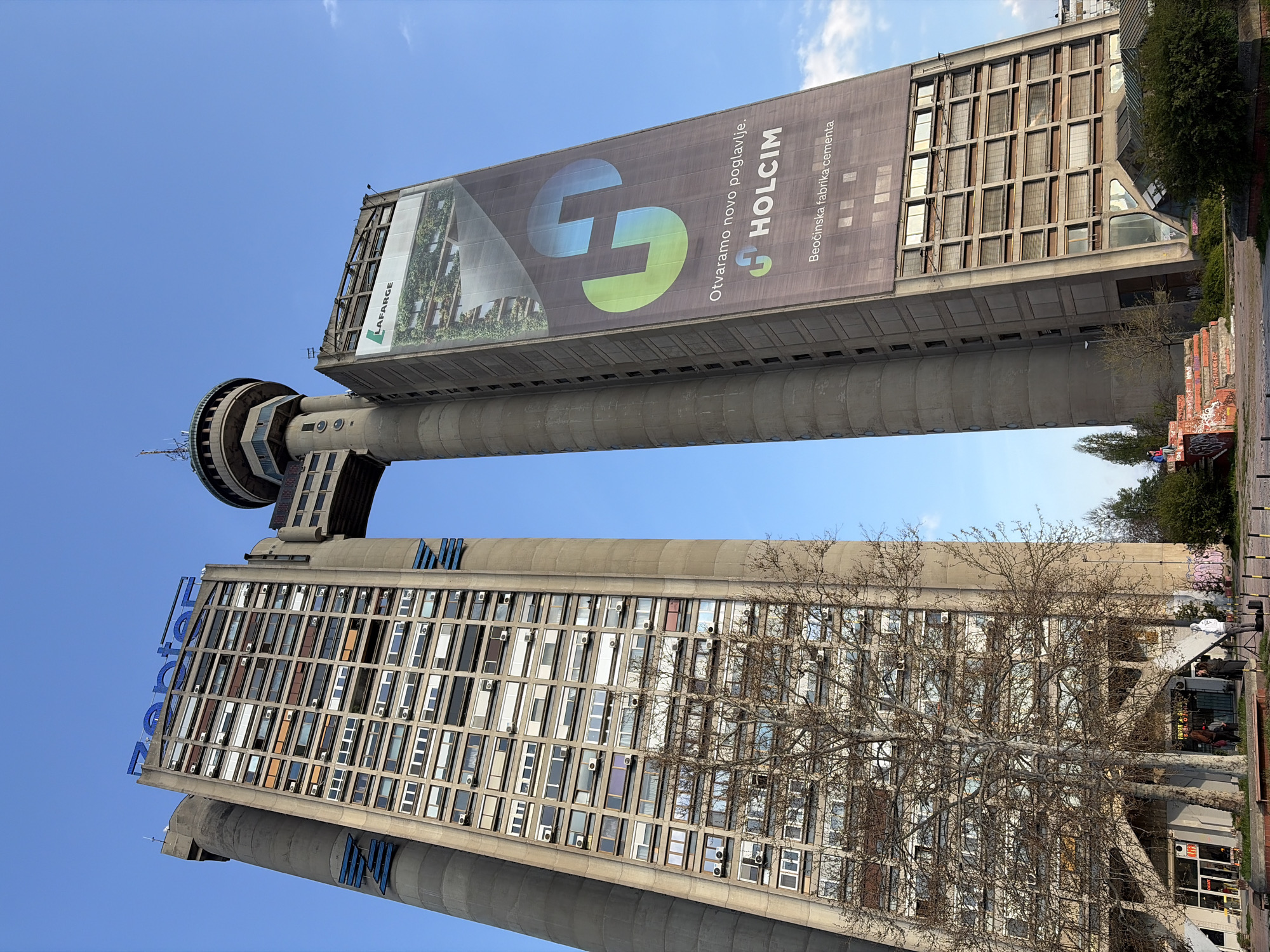 The image shows the Genex Tower, a high-rise building in Belgrade, Serbia. The building consists of two concrete towers connected by a central core and a rotating restaurant at the top. The towers are covered with windows. A large advertisement for Holcim Cement is visible on the upper tower. The sky is blue and clear. A bare tree is visible in the foreground. People are sitting on a nearby platform.