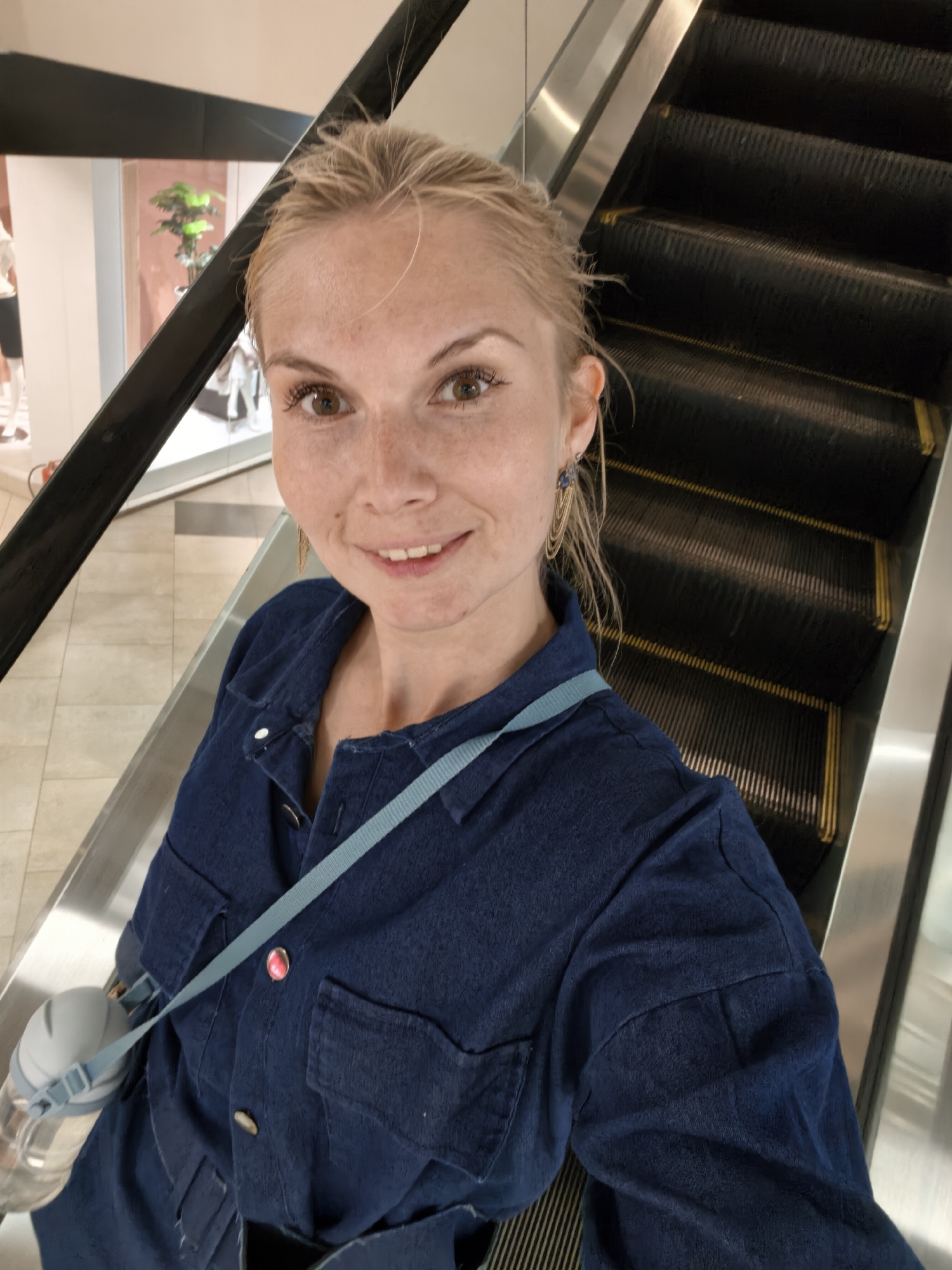 A young blonde woman is taking a selfie while standing on an escalator. She is wearing a dark blue denim shirt with a light blue strap across her chest, and she has a water bottle attached to the strap. Her hair is pulled back, and she is smiling at the camera. The escalator is dark gray with yellow safety lines. In the background, there is a store display with mannequins and a plant. The flooring appears to be tiled. The scene is likely indoors, possibly in a shopping mall or department store.