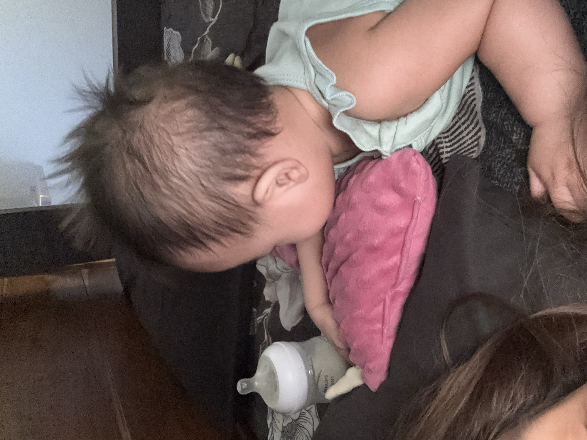 A baby is lying on its stomach on a dark grey surface with black and white striped fabric visible underneath. The baby is wearing a light green sleeveless top with a ruffled neckline. A pink pillow is positioned beneath the baby's head and torso. To the left of the baby is a clear baby bottle with a white nipple, containing a light-colored liquid. The baby's hair is dark and appears to be standing up in some places. The scene is indoors, and no specific location in West Jakarta, Indonesia can be determined from the image.