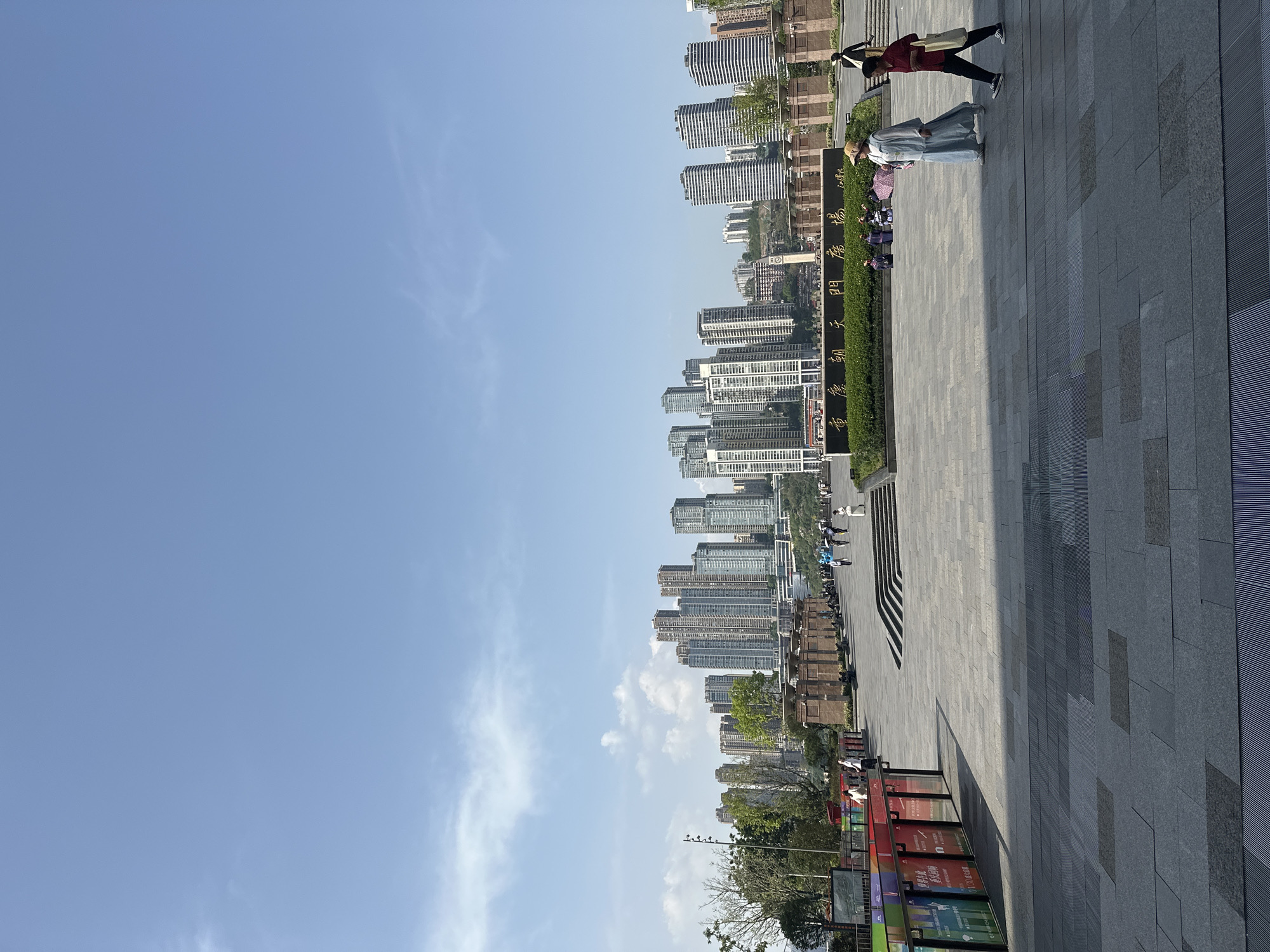 The image shows an outdoor scene in Yuzhong District, Chongqing, China, on a sunny day with a clear blue sky. A group of tall buildings are visible in the background. Closer to the foreground, there is a wide pedestrian area with gray pavement. People are walking, and some are dressed in traditional Chinese clothing. There are green hedges and memorial plaques. A row of colorful advertising panels stand near the edge of the pavement.