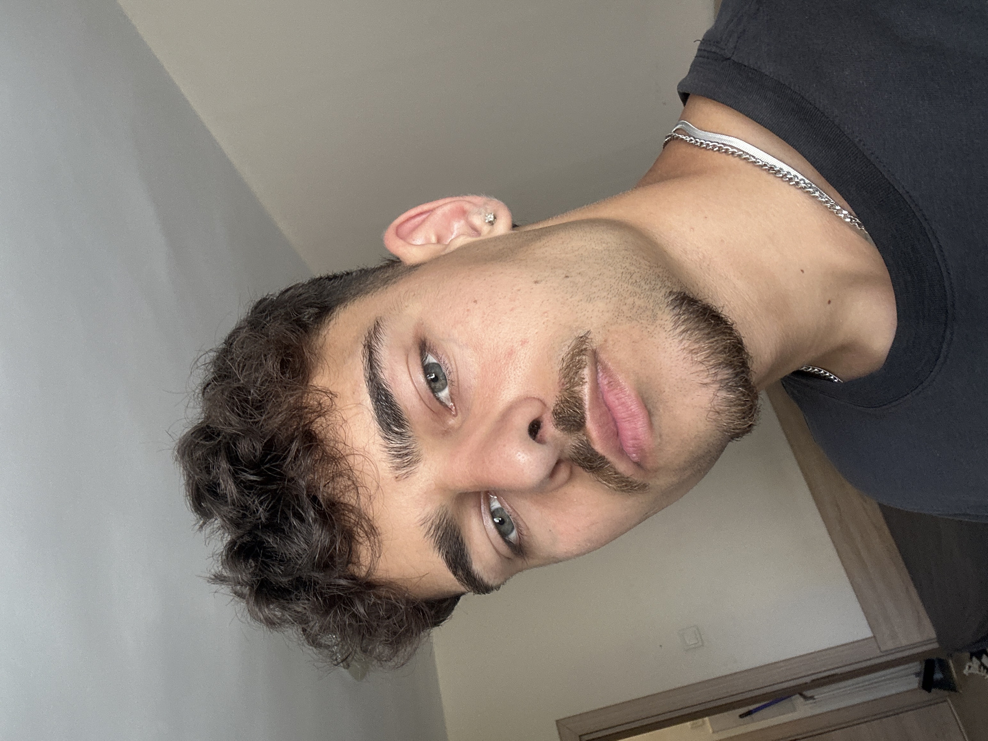 A young man with dark curly hair, light eyes, and facial hair is posing for a close-up, self-portrait photograph. He is wearing a dark T-shirt and a silver chain necklace. He appears to have an earring. The background includes a light-colored wall and a partially visible doorway or cabinet. The photo appears to have been taken indoors, likely in an apartment or a room in a residential building. There is no information that the photo has been taken in the city of Sofia, Bulgaria.