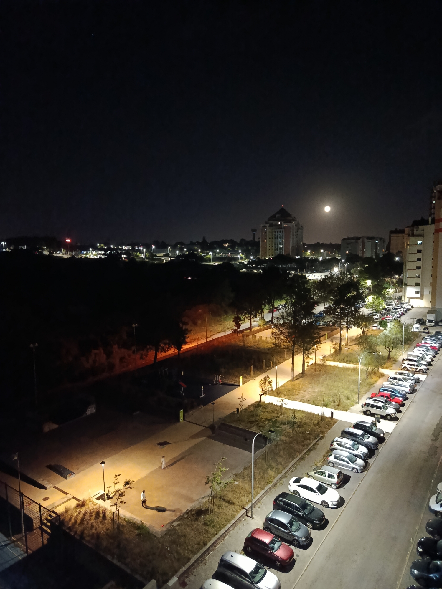 The image captures a nighttime scene in Algueirão-Mem Martins, Portugal, with a full moon prominently visible in the sky. The foreground features a parking lot filled with numerous cars of various colors and models, parked in rows alongside a street. Several streetlights illuminate the area, casting long shadows. In the middle ground, a park or recreational area is visible, with pathways, trees, and playground equipment. Two individuals can be seen walking in the park. Beyond the park, a cityscape unfolds, characterized by buildings of different heights and architectural styles. One building, a prominent, tall structure with a pyramidal top, stands out against the dark sky. The overall atmosphere is tranquil, with the urban landscape illuminated by artificial lights and the natural glow of the moon.