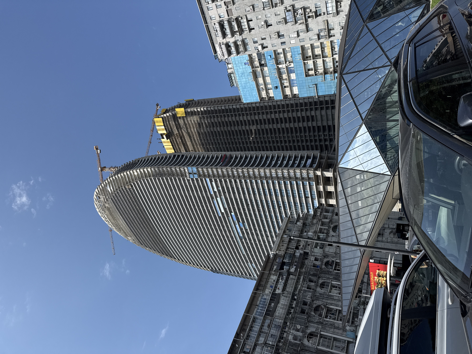 An eye-level, exterior shot captures a bright, sunny day in an urban setting with a clear blue sky and scattered white clouds. The foreground is dominated by the hoods of several parked cars, with a dark SUV on the right and a lighter-colored vehicle to its left. Behind the cars, a striking, modern glass-paneled structure with a geometric, faceted design is visible.
The middle ground features several buildings. To the left, a tall, curved skyscraper, seemingly under construction, rises dramatically. Its facade is composed of horizontal white louvers and vertical structural elements, with a crane visible at its peak. Next to it, another, taller skyscraper is also under construction, its concrete frame exposed, adorned with yellow scaffolding at the top and another crane. To the right of these new structures, an older, grey apartment building with numerous balconies and visible air conditioning units stands. Further right, a modern building with a bright blue glass facade is partially visible, with balconies and windows interspersed.
The background consists of a clear blue sky. The overall scene depicts a city undergoing development, with contrasting architectural styles of old and new buildings.