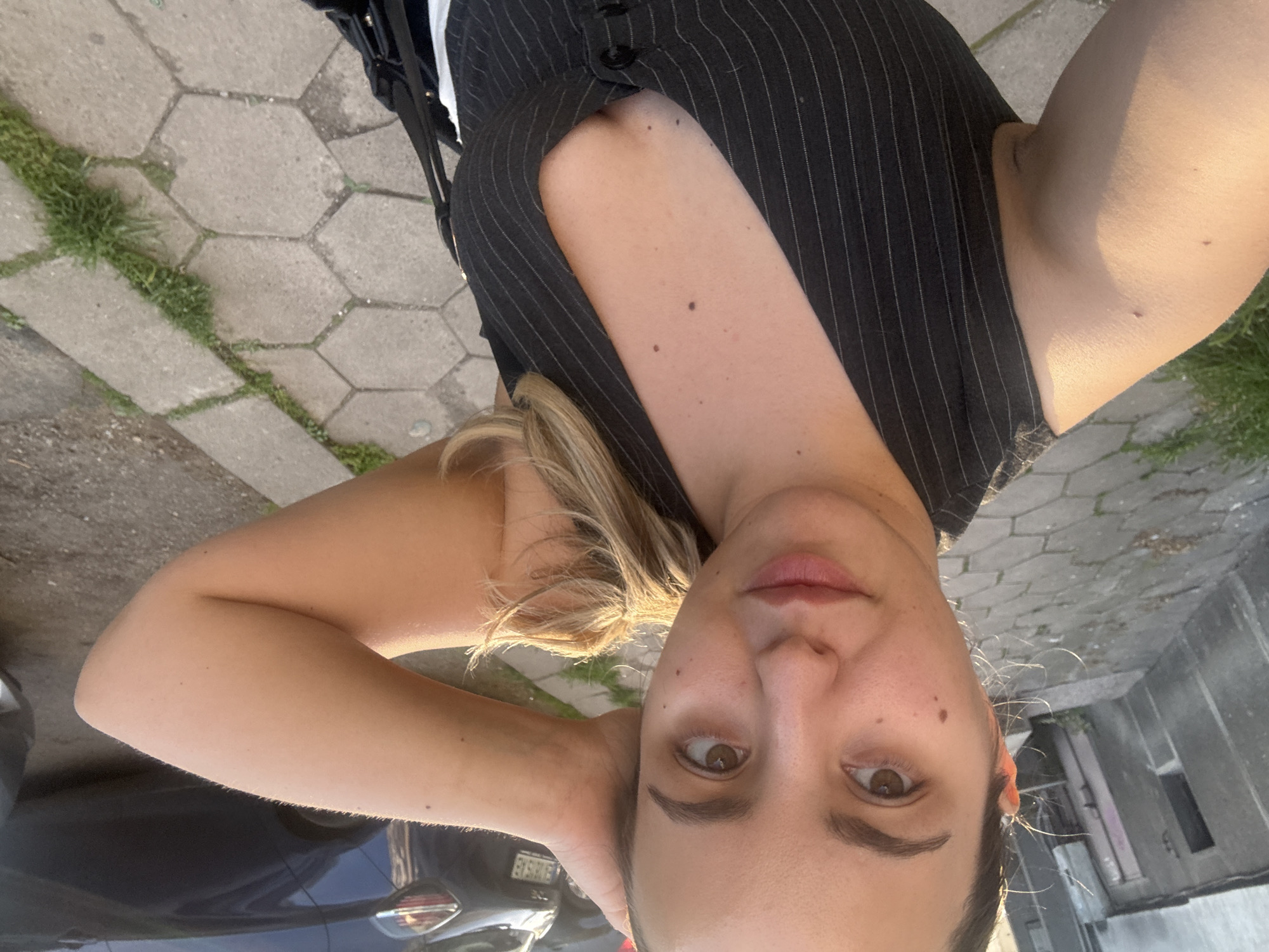 A selfie, taken at a high angle, shows a young woman with fair skin, light brown eyes, and blonde hair, wearing a black pinstripe vest. She is standing on a hexagonal-paved sidewalk in an urban area. Some grass grows between the pavement stones. In the bottom left, part of a dark blue car is visible. A building is visible in the upper right of the frame. The picture was taken outdoors during the day. I am unable to locate this image in Sofia, Bulgaria, as there is no discernible information within the image to make that conclusion.