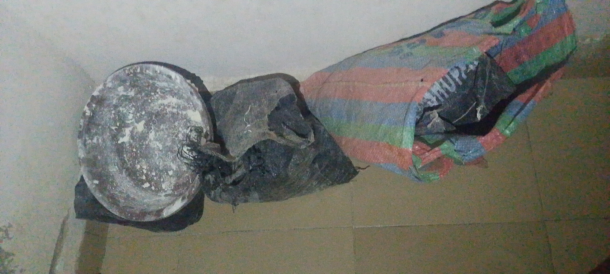 In Jauro Yinu, Nigeria, a scene depicts a collection of items placed on a tiled floor. To the left, a round, flat object, possibly a lid or tray, is visible. It is speckled with white residue. Next to it, a dark, irregularly shaped object appears to be a bundle of material, possibly fabric or sacks, with some fraying. Extending from this is a large, striped bag with horizontal bands of red, blue, green, and possibly yellow. The bag is open, revealing dark contents within, and appears to be made of a woven material. The background is a plain, light-colored wall. The overall impression is one of discarded or stored items. There are no people present in the image.