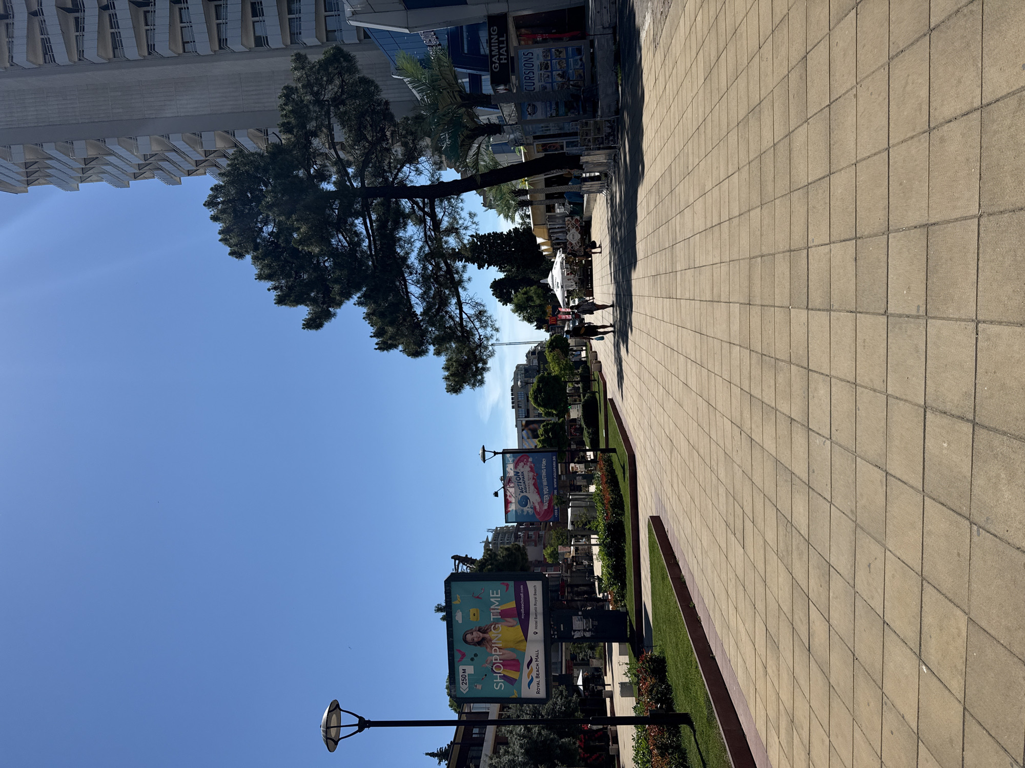 The image depicts a sunny day scene in Nesebar, Bulgaria, likely on a pedestrian walkway or promenade. In the foreground, a wide tiled pathway stretches into the distance, bordered by manicured green lawns and flowerbeds. A lamppost stands to the left, and further down, a large billboard advertises "Shopping Time" at "Royal Beach Mall." To the right, a modern, multi-story building with balconies dominates the background, with a large pine tree in the middle ground casting shadows. Other buildings, shops, and a few palm trees are visible further along the walkway. Two individuals are walking away from the camera on the right side of the pathway, and a few more figures are scattered further down. Signs for "Action Aquapark" and "Gaming Hall" are also visible. The sky is clear and blue, indicating fair weather.