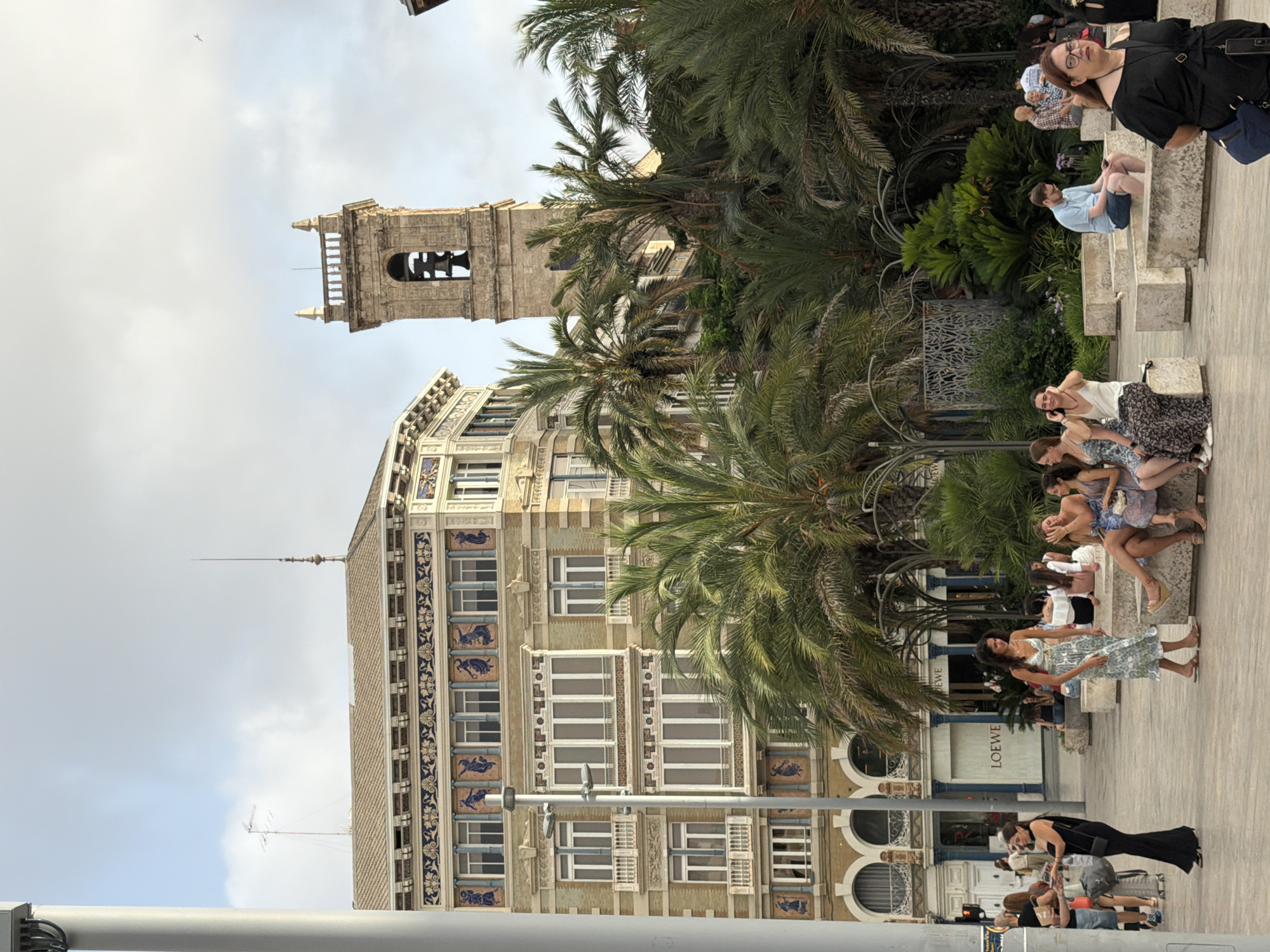 The image captures a sunny outdoor scene in an urban environment, likely a public square. A multi-story building with ornate architectural details, featuring decorative friezes and numerous windows, dominates the background. To the right of the building, a tall, ancient-looking stone tower with a bell is visible, suggesting a historical or religious site. Lush palm trees and other greenery are scattered throughout the scene, creating a tropical or Mediterranean ambiance.
In the foreground and mid-ground, several people are engaged in various activities. A woman in a black formal dress is walking to the left, pulling a suitcase. Others are seated on low, rectangular stone benches, some appearing to be in conversation while others are looking at their phones. A group of young women in casual summer attire are also seated, one of whom is on her phone. A man in a blue shirt and shorts is seated on a bench, also focused on his phone. Another woman, wearing glasses and a black top, stands to the right, looking towards the viewer. The overall impression is a lively public space where people are enjoying a pleasant day outdoors. The presence of a "LOEWE" storefront indicates a commercial area within the city.