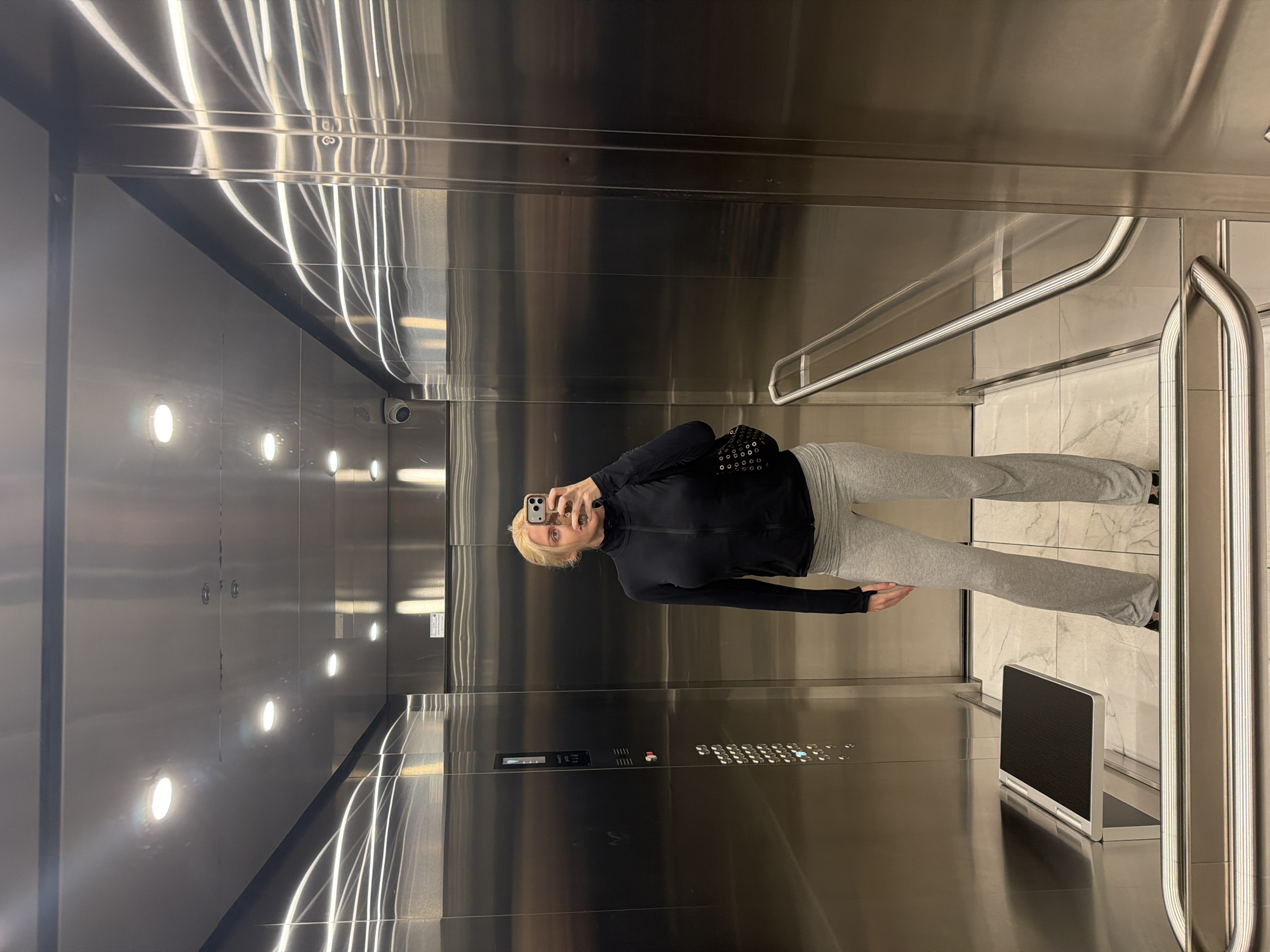 A woman is taking a selfie in an elevator with stainless steel walls. The elevator has a control panel with buttons and a small screen on the right side. There is a handrail on the right side. The elevator interior is illuminated by rows of lights on the wall. The woman is wearing a black top and gray sweatpants. She is holding a smartphone in her left hand to take the picture. The location is not discernible from the image. There is no evidence to place the location in Belgrade, Serbia.