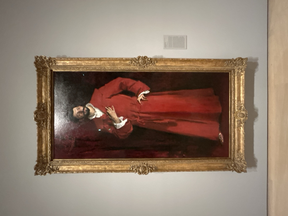 The image displays a large oil-on-canvas portrait titled "Portrait of a Gentleman in Red" by El Greco (Doménikos Theotokópoulos), painted circa 1570-1575, exhibited at the Norton Simon Museum in Pasadena, Los Angeles County, California, United States of America.
The vertical painting depicts a standing male figure, full-length, facing slightly left with his head turned to look towards the right. He possesses a dark beard and mustache. The man is dressed in a long, flowing, deep crimson-red robe, cinched at the waist with a matching sash ending in tassels. A white ruffled shirt with an open collar and cuffs is visible beneath the robe. His right hand rests flat against his upper chest, while his left hand is positioned on his hip with fingers extended. Light-colored slippers are visible at the bottom of the robe. The background of the portrait is a dark, monochromatic reddish-brown.
The painting is enclosed within an elaborate, gilded wooden frame featuring ornate carvings and classical embellishments. It hangs centrally on a plain, light grey wall. To the right of the painting, a rectangular white information plaque with black text provides details about the artwork. The floor visible at the base of the wall is constructed of light-colored wood. Reflective glare from an external light source is visible on the painting's surface. No individuals are present within the captured scene.