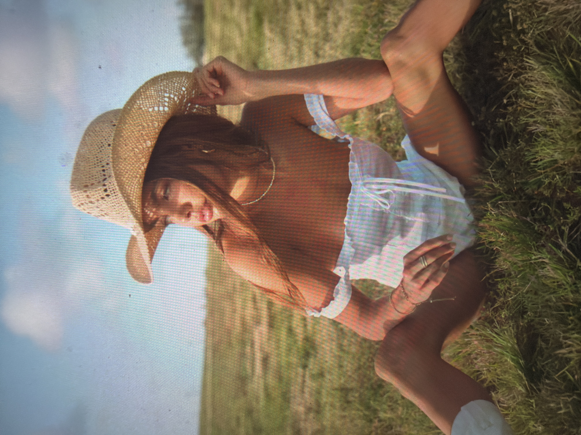 The image depicts a young female subject seated in an expansive, sunlit grassy field under a blue sky with scattered white clouds. The subject, centrally framed, wears a light-colored, possibly white or pale blue, off-the-shoulder top with a ruffled neckline and a tied drawstring front closure. She wears a woven straw cowboy hat. Her long, dark brown hair falls over her shoulders and partially obscures her face. A thin chain necklace is visible, along with a bracelet on her left wrist and a ring on her left ring finger. Her left hand is positioned near the brim of her hat, while her right arm rests on her bent knee. Her legs are spread, with her bare skin visible above her knees. The subject's gaze is directed towards the upper right of the frame. The immediate foreground and mid-ground consist of green and dry grass, extending to a discernible horizon line where it meets the sky. The lighting indicates direct, warm sunlight, casting soft shadows. The image exhibits visible pixelation, suggesting it is a photograph of a digital screen. The visual content of the image does not contain any identifiable landmarks or features to confirm the location as Shumen, Bulgaria.