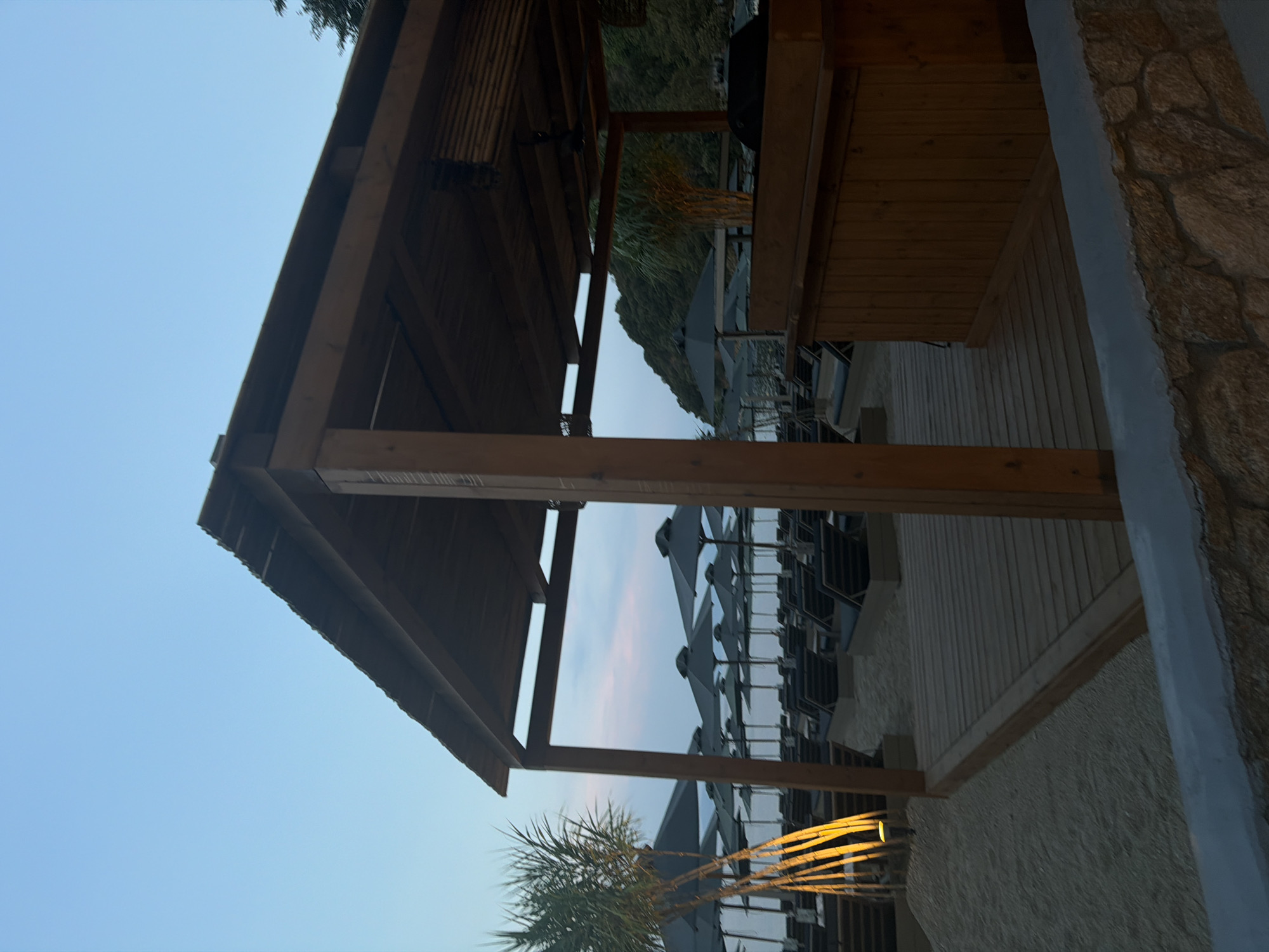 The image depicts an outdoor bar area on a sandy beach in Sithonia Municipal Unit, Greece, during twilight. The structure is made of wood, with a sloped roof and exposed beams. In the foreground, a wooden deck area transitions to sand. Behind the bar, rows of empty lounge chairs with dark umbrellas are set up on the beach, facing the sea. A tropical-looking plant with yellow uplighting is visible to the left of the bar. In the background, a tree-covered hillside slopes down towards the water. The sky is a clear, pale blue with hints of pink from the setting sun. There are no people visible in the image.