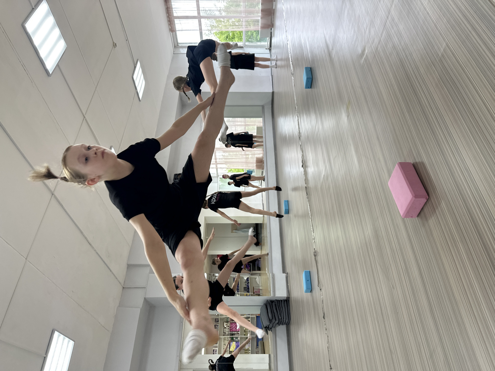 In a dance studio in Votkinsk, Russia, a young female dancer, her blonde hair pulled into a high ponytail, is captured mid-air performing a split jump. She is wearing a black leotard and white socks. Her body is oriented horizontally, with her legs extended wide apart, demonstrating a high level of flexibility and training. The studio environment includes a mirrored wall reflecting other dancers in similar attire, as well as shelves filled with trophies. The floor is a light wood laminate, and several colorful yoga blocks are scattered on the floor, one pink and two blue, suggesting training equipment. The lighting in the room is provided by fluorescent ceiling panels. The overall atmosphere suggests a practice or rehearsal session for young dancers.