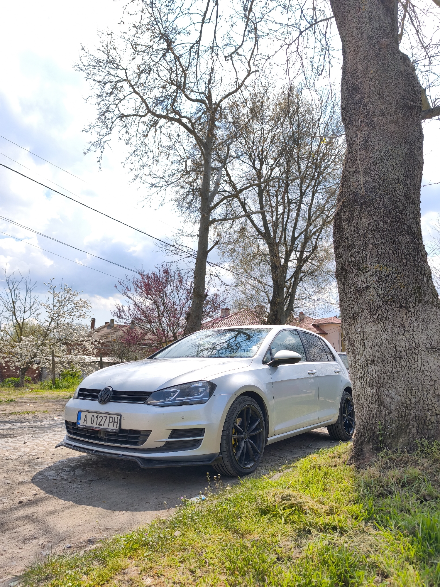 A silver Volkswagen Golf sits parked on a dirt road adjacent to a grassy area. The car has black wheels and a Bulgarian license plate reading "A 0127 PH." A large tree trunk is immediately to the right of the car. Behind the car and to the left, trees with budding leaves and flowering branches are visible in the background. Beyond these trees, glimpses of houses with red-tiled roofs can be seen. Utility lines stretch across the upper left portion of the image. The sky is partly cloudy. This image appears to be located near Rusokastro, Bulgaria.