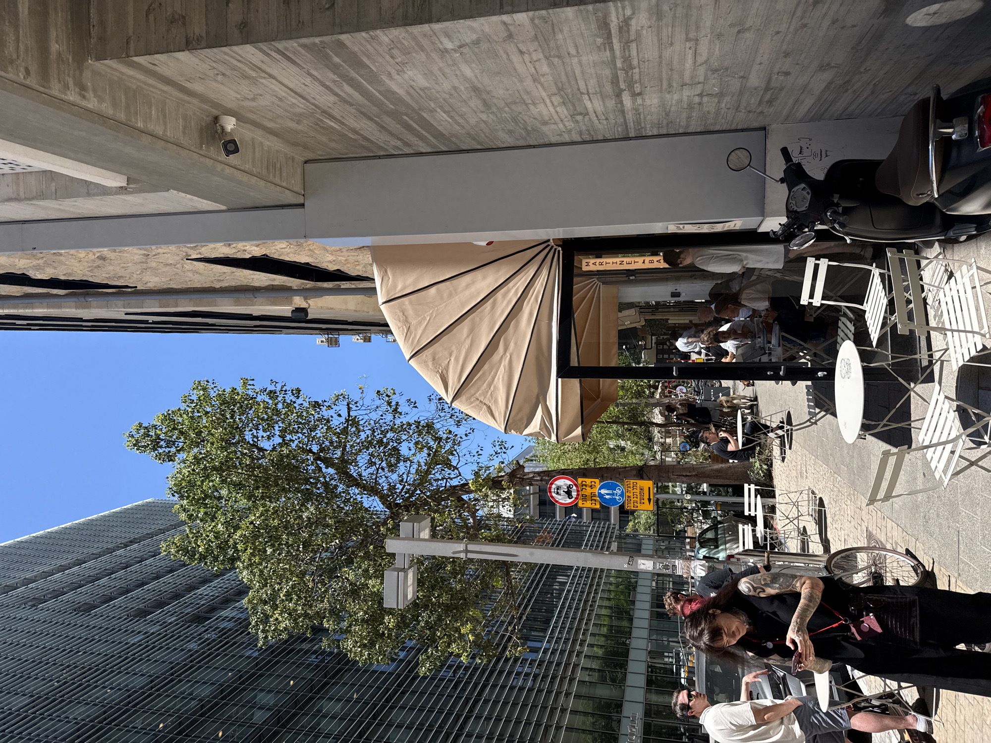 This is a street-level, medium-shot photograph captured on a bright, sunny day in Tel-Aviv, Israel. The scene depicts a bustling urban environment with an emphasis on an outdoor cafe area.
Dominating the left side of the frame is a modern, tall building with a glass facade, characterized by horizontal louvers, indicative of its contemporary architectural style. To the right, a building with a textured, concrete exterior and stone accents presents a stark contrast. A light-colored awning extends from this building, covering an outdoor seating area for what appears to be a cafe or restaurant.
Several people are visible in the image. In the foreground, a woman with dark hair and tattoos on her arms is standing, looking at her smartphone. She is wearing a black vest and pants, with a dark crossbody bag. To her left, a man in a white t-shirt and grey shorts is leaning against a car. Further into the cafe area, other individuals are seated at tables, engaged in conversation or dining. A black scooter is parked in the foreground on the right.
Street furniture, including white metal tables and chairs, is arranged on the sidewalk, suggesting a pedestrian-friendly zone. Traffic signs are visible on a pole, including one with a red circle and a diagonal line over a motorcycle, indicating a prohibition, and others with symbols for pedestrians and bicycles. The ground is paved with interlocking blocks, typical of urban sidewalks.
The overall atmosphere is one of a lively city day, with people enjoying the outdoor space amidst the urban landscape. The presence of trees adds a touch of greenery to the otherwise concrete and glass environment.