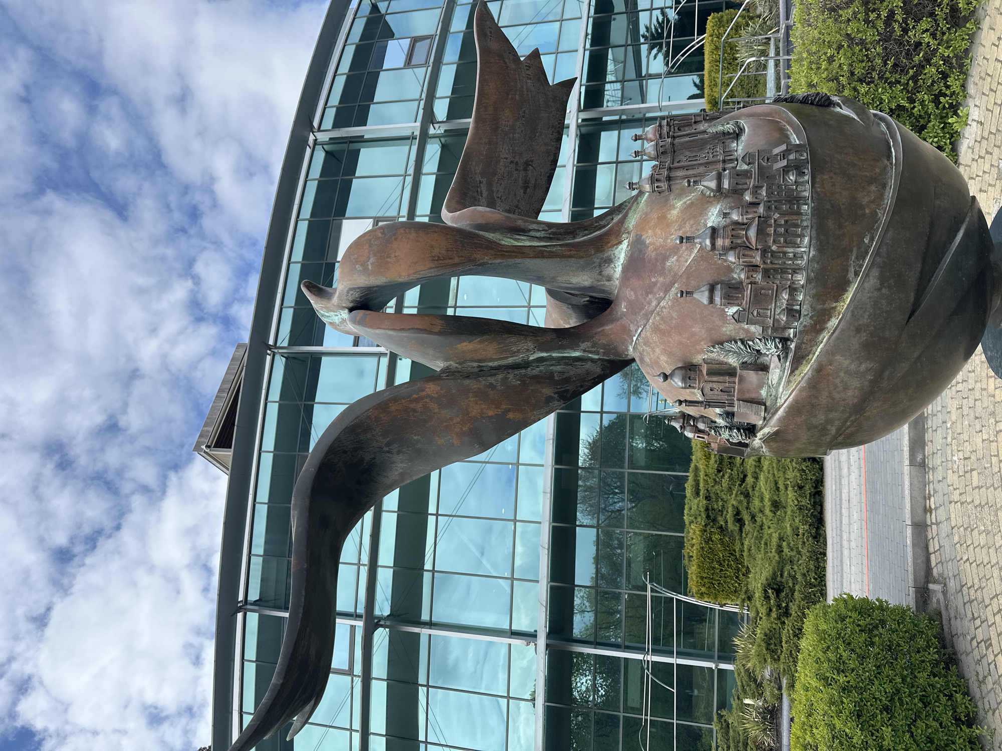 A striking bronze sculpture stands prominently, depicting an intricate combination of forms. The sculpture features a large, spherical or globe-like element adorned with numerous miniature architectural structures, resembling a densely packed cityscape of historical buildings, complete with domes, spires, and detailed facades. This "city" appears to be integrated with or carried by flowing, abstract bronze shapes that evoke the sense of wings, sails, or ribbons in motion, suggesting flight or dynamic movement. The bronze has a weathered patina, showing shades of brown, green, and black from oxidation.
Behind the sculpture rises a modern building with a distinctive curved glass facade. The glass panels, framed by light-colored structural elements, reflect the bright, cloudy sky and the surrounding greenery. Lush green bushes and manicured landscaping are visible at the base of the sculpture and along the building, adding a natural contrast to the metallic and glass structures. A paved area or pathway is visible to the right and bottom of the frame, constructed from light-colored pavers.
The scene takes place during the daytime under a sky filled with scattered white clouds. There is no visible text or human activity. The overall impression is one of architectural beauty and artistic expression, blending historical representation with modern design in an outdoor setting in Kyiv, Ukraine.
