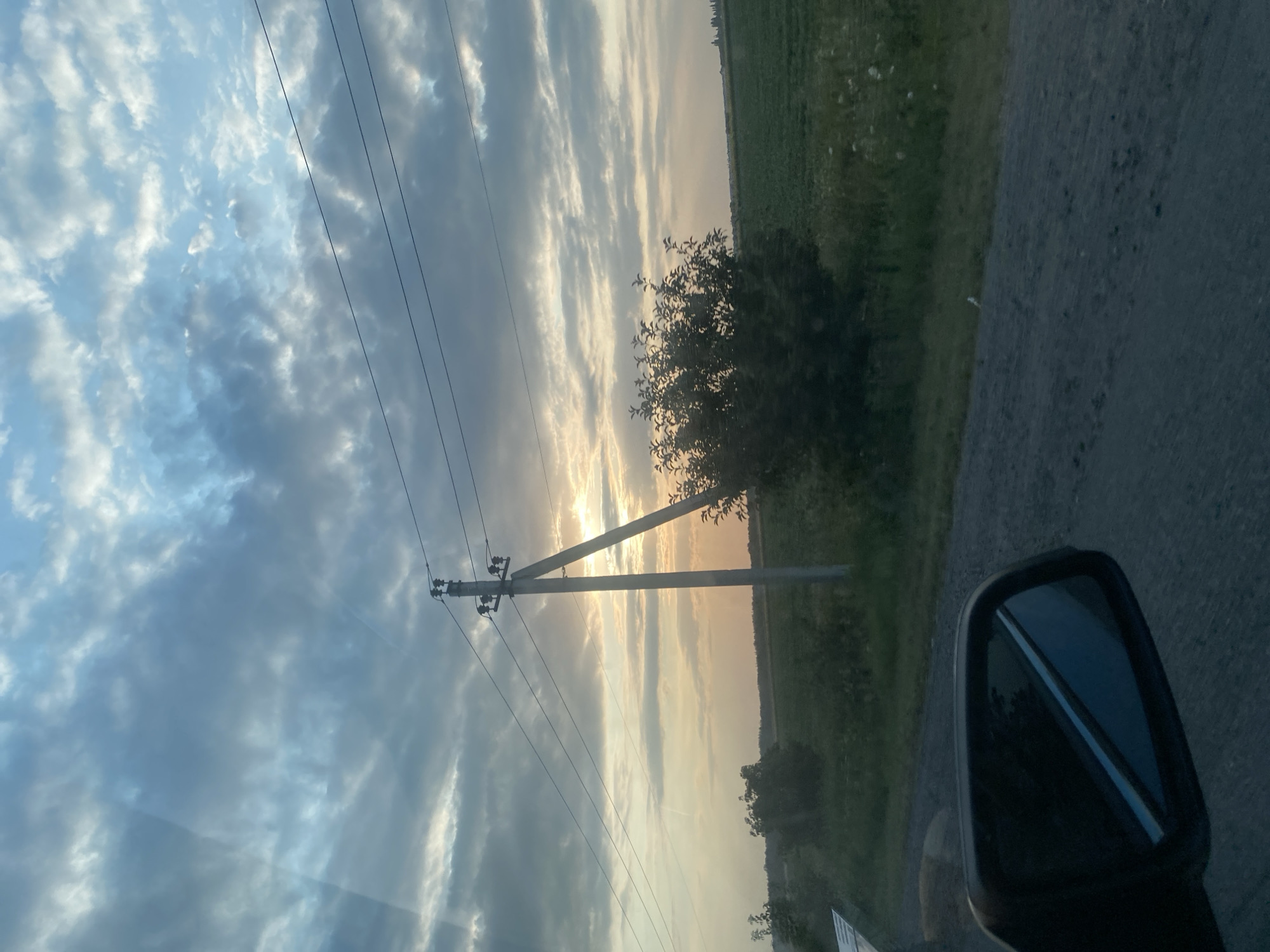 The image depicts a sunset view of a rural landscape, likely near Pishchanka, Ukraine. In the foreground, a car's side mirror is visible on the left, reflecting the sky. The road appears to be unpaved. A utility pole with wires stands prominently in the middle ground, supporting electrical lines that stretch across the frame. Beyond the road and pole, a green field extends towards the horizon, with a line of trees and bushes bordering it. The sky is filled with dramatic clouds, illuminated by the setting sun, which is partially obscured by the clouds, casting a warm, golden light. The overall scene conveys a sense of twilight in a rural setting.