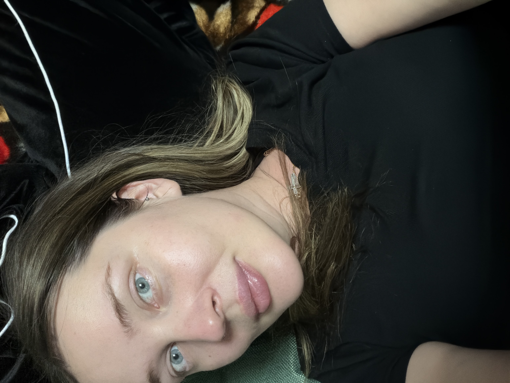 A woman with light skin, blue eyes, and light brown hair with blonde highlights is lying down. She is wearing a black shirt and a gold cross necklace. She appears to be indoors. The location is unknown.