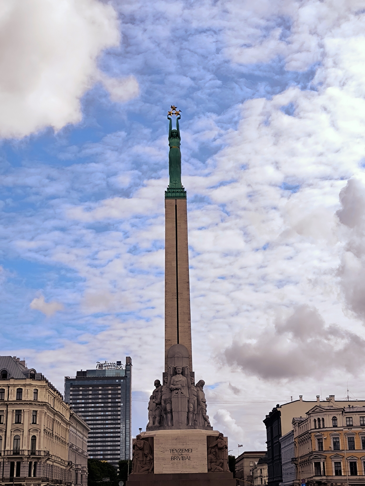 The image captures the Monument of Freedom in Riga, Latvia, a prominent landmark in the city. The monument stands tall against a backdrop of a blue sky with scattered clouds, some of which are dense and white, while others are lighter and more wispy. At the apex of the monument, a green copper statue of a woman, known as Milda, holds three golden stars above her head. Below her, the obelisk is adorned with sculptures depicting various figures, including what appear to be soldiers and other individuals. The base of the monument features inscriptions in Latvian, one of which reads "Tēvzemei un Brīvībai" (For Fatherland and Freedom). To the left of the monument, a tall, modern glass-fronted building with the "Radisson" logo is visible, contrasting with the classical architecture of the older buildings lining the street in the foreground. These older buildings exhibit ornate facades with balconies and numerous windows. To the right, more traditional European architecture lines the street, with buildings featuring pitched roofs and decorative elements. The scene appears to be a public square or plaza, given the presence of the monument and surrounding urban architecture. The lighting suggests it is daytime, with soft light illuminating the scene.