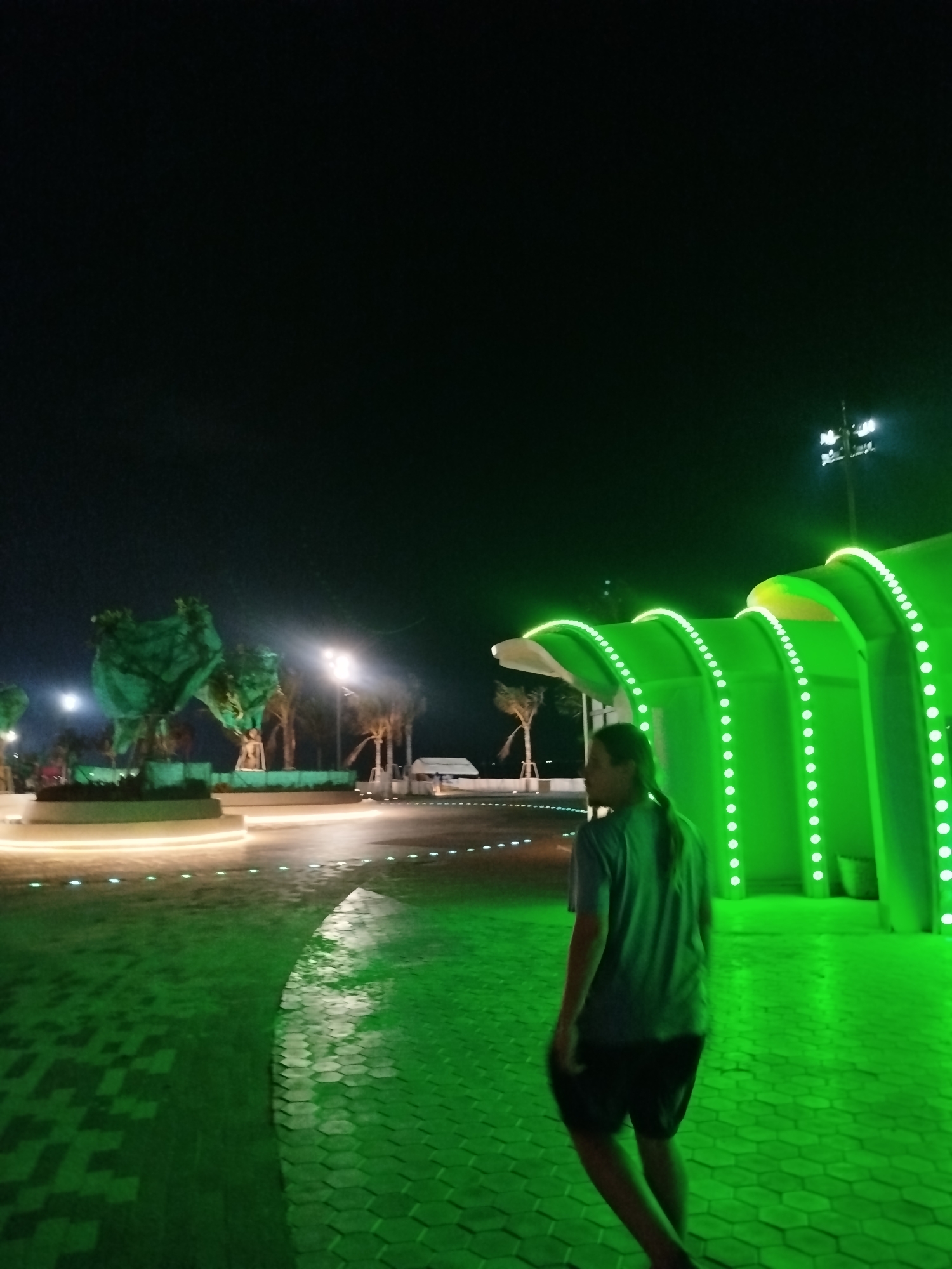 A person with long hair pulled back in a ponytail is walking on a paved walkway at night. The walkway is lit with green light, and there are green-lit structures and trees in the background. In the distance, there is a white tent. There are lights to the right of the frame that resemble stadium lights. Based on the available context, the image shows a scene at night in Ho Chi Minh City, Vietnam.