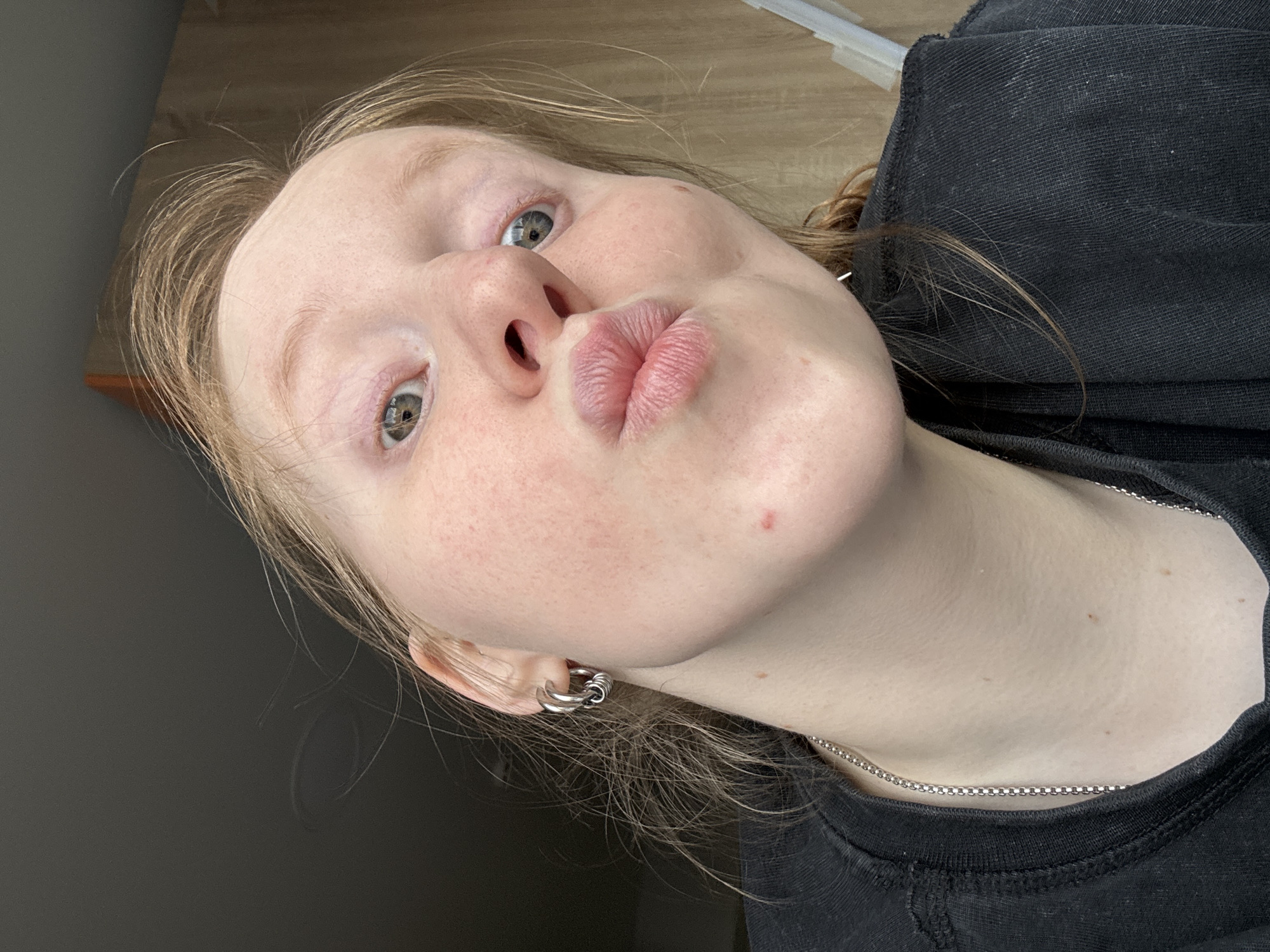 A close-up, upward-angled selfie depicts a young woman with fair skin and light blonde hair. Her lips are pursed in a kissing expression. She has light-colored eyes and freckles are visible on her face. She is wearing a black top and a silver chain necklace. A silver earring is in her left ear. The background is a mix of light and dark tones, indicating an indoor setting. The location (Perm, Russia) cannot be confirmed by the image.