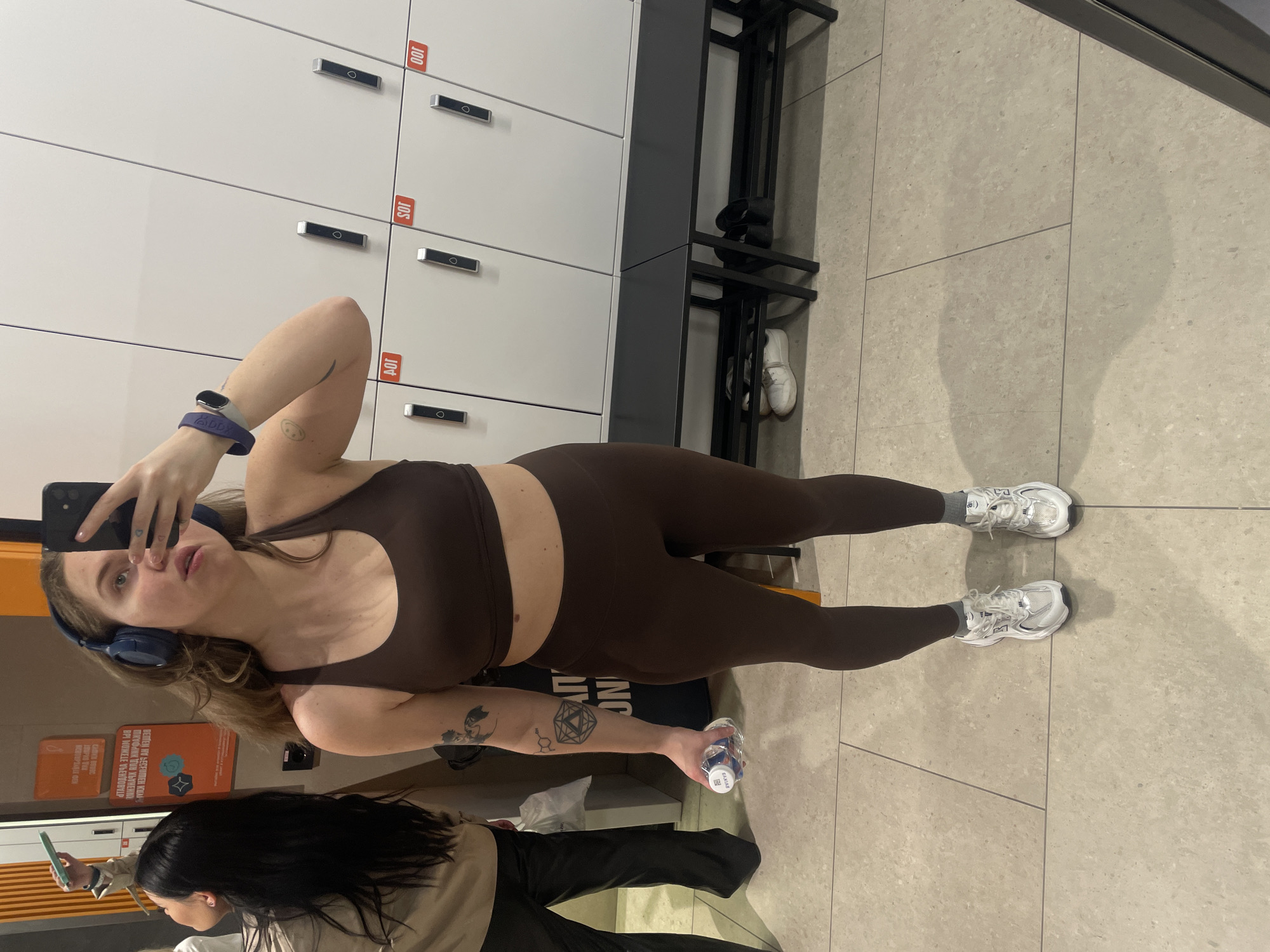 The image is a selfie taken indoors, likely in a gym or fitness center, showing a woman in exercise attire. She is wearing a brown sports bra and leggings set, along with white sneakers. A blue fitness tracker is visible on her wrist. She is holding a smartphone in her hand and taking the selfie, while also holding a water bottle in her other hand. She has headphones around her neck. Behind her are white lockers with red labels and a metal bench with shoes placed on it. Another woman with long black hair is partially visible in the lower part of the frame, wearing a beige jacket and dark pants, possibly interacting with a phone. There is also an orange poster with Russian text on the wall behind her. The floor is tiled with a light grey pattern. Based on the poster written in Russian, it can be inferred that the location is within the city of Moscow, Russia.