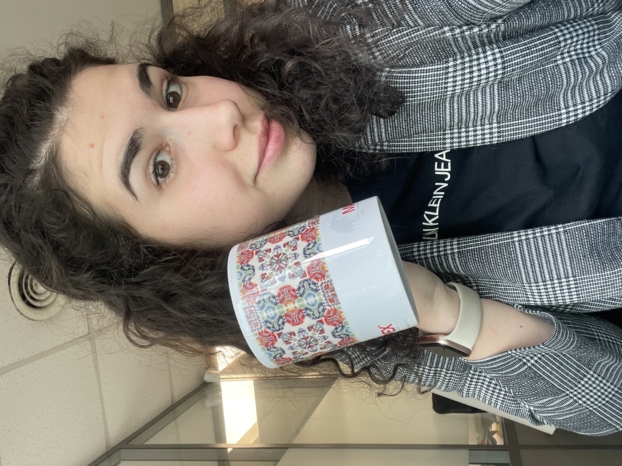 The image is a selfie of a woman holding a patterned mug. She has curly dark hair, fair skin, and is wearing a black t-shirt with the words "CALVIN KLEIN JEANS" partially visible, and a black and white plaid blazer. She's also wearing a light-colored smartwatch.
The mug she is holding is white with a colorful embroidered-like pattern around it. The background appears to be an indoor office setting, with a visible window and a ceiling with a vent. Given the pattern on the mug, which is a typical Bulgarian embroidery and textile, and that the patterns shown on the mug are traditionally from the area of Sofia, Bulgaria, it's plausible that the photo was taken in Sofia, Bulgaria.